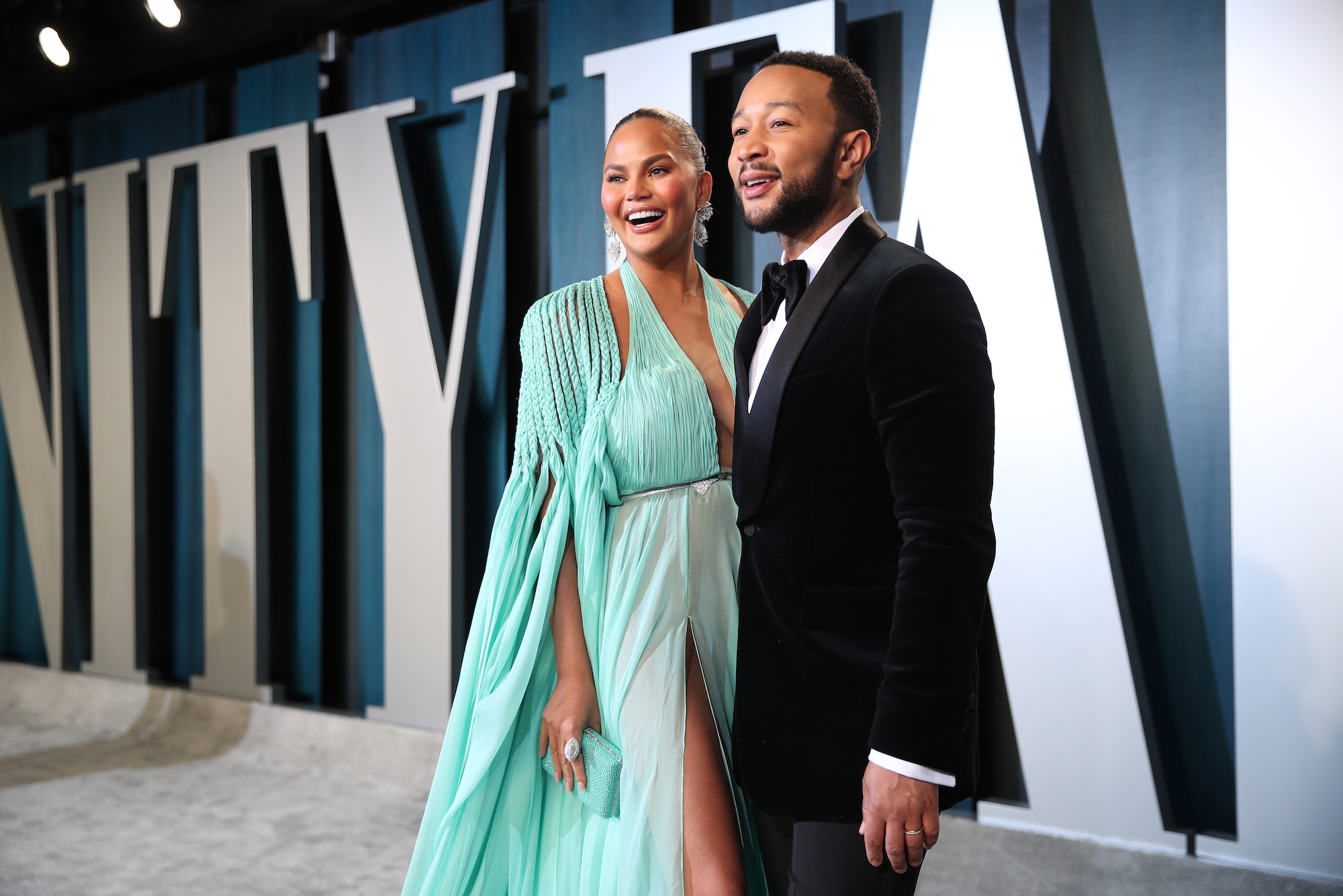 (L-R) Chrissy Teigen and John Legend smiling in front of a blue and white background on an awards show carpet