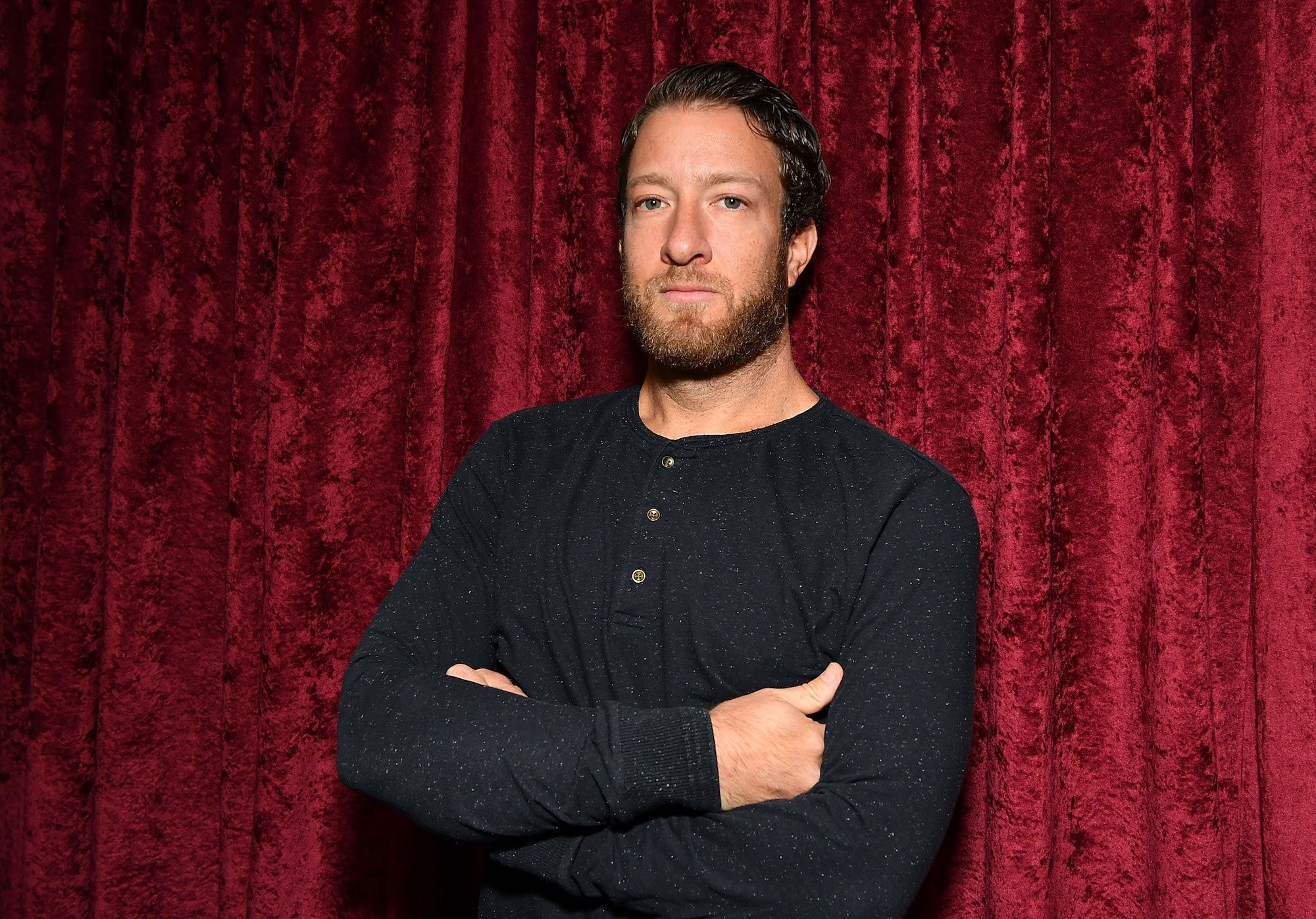 Dave Portnoy standing with his arms crossed in front of a red velvet curtain