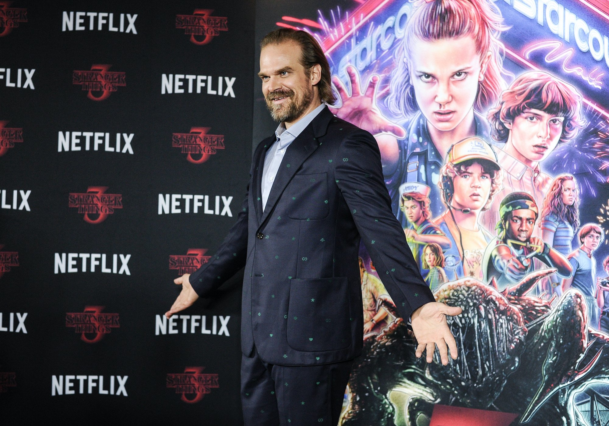 David Harbour smiling, arms outstretched, in front of a 'Stranger Things' poster