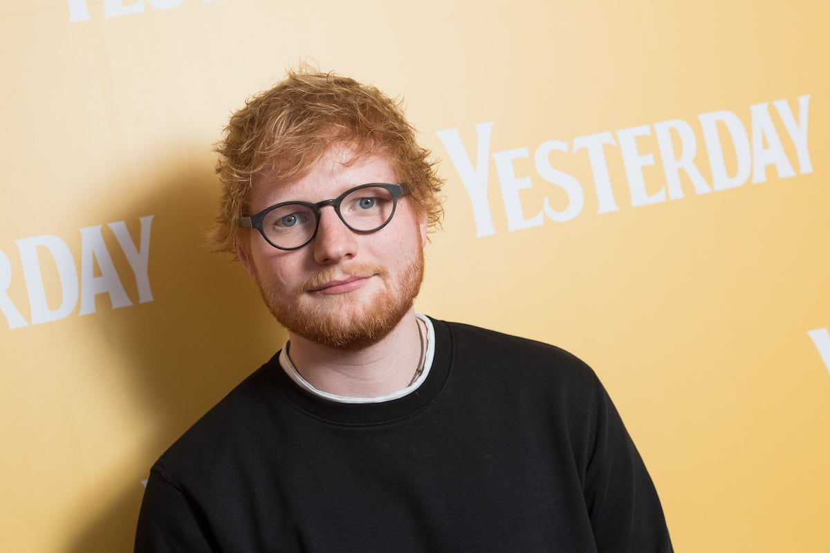 Ed Sheeran looks at the camera wearing a black shirt against a soft yellow background