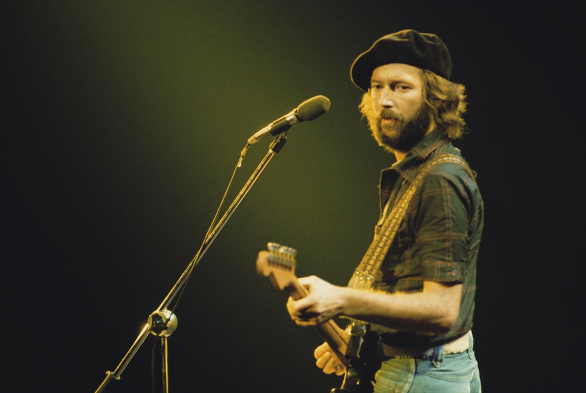 British guitarist Eric Clapton performing on stage during his US tour, July 1975. He's on stage holding a guitar and staring to his left, wearing denims, a black shirt and a black hat. 