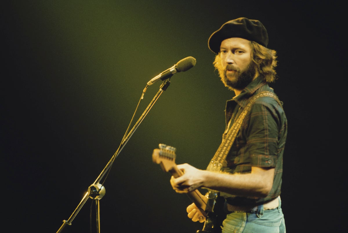 British guitarist Eric Clapton performing on stage during his US tour, July 1975. He's on stage holding a guitar and staring to his left, wearing denims, a black shirt and a black hat.