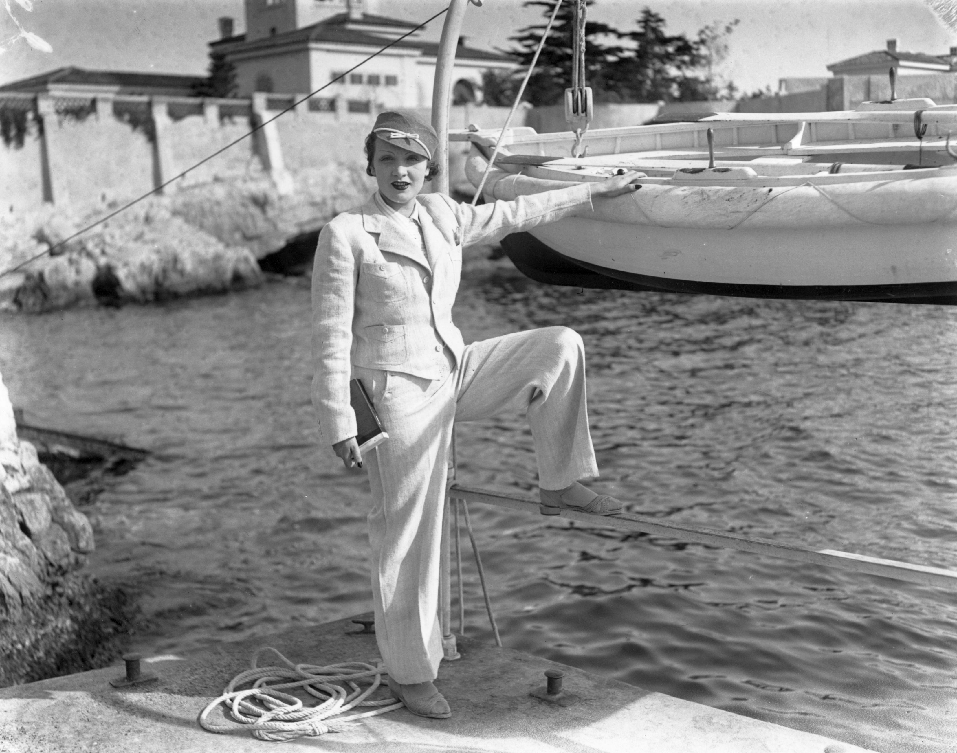 Marlene Dietrich in a pantsuit standing next to a waterfront, in black and white