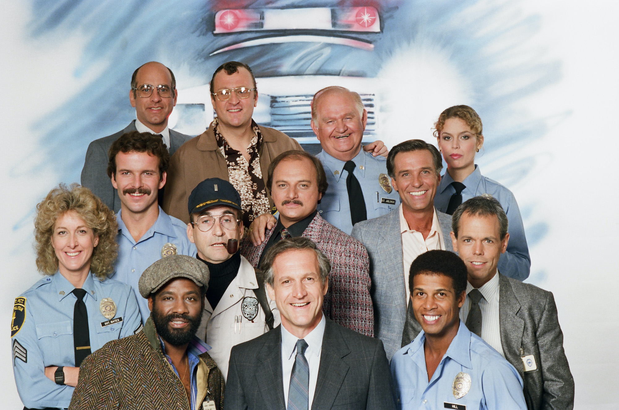 Hill Street Blues season 7 cast smiling in front of a backdrop featuring an illustration of a police car