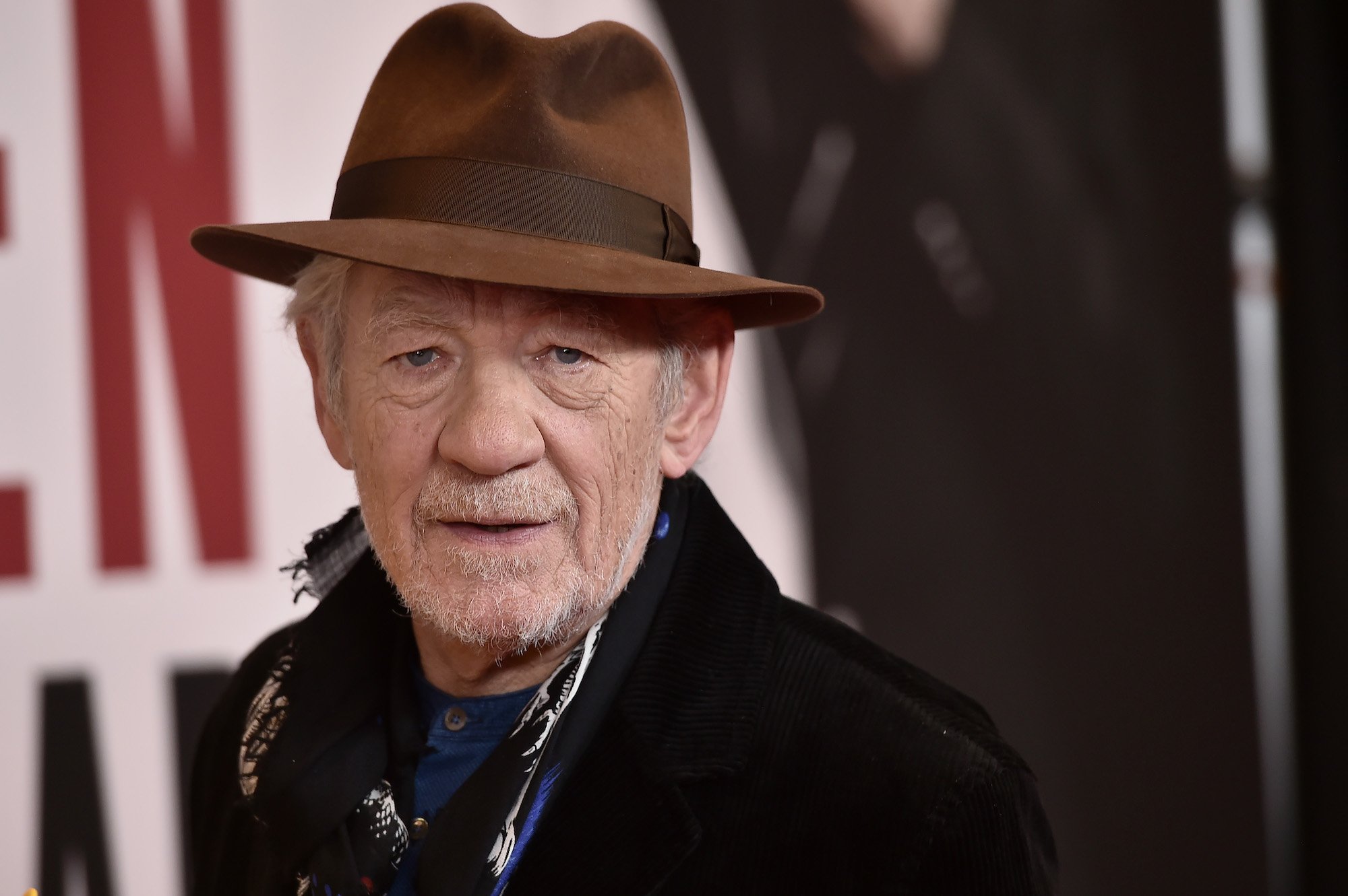 Sir Ian McKellen looking at the camera in front of a blurred background, wearing a brown hat