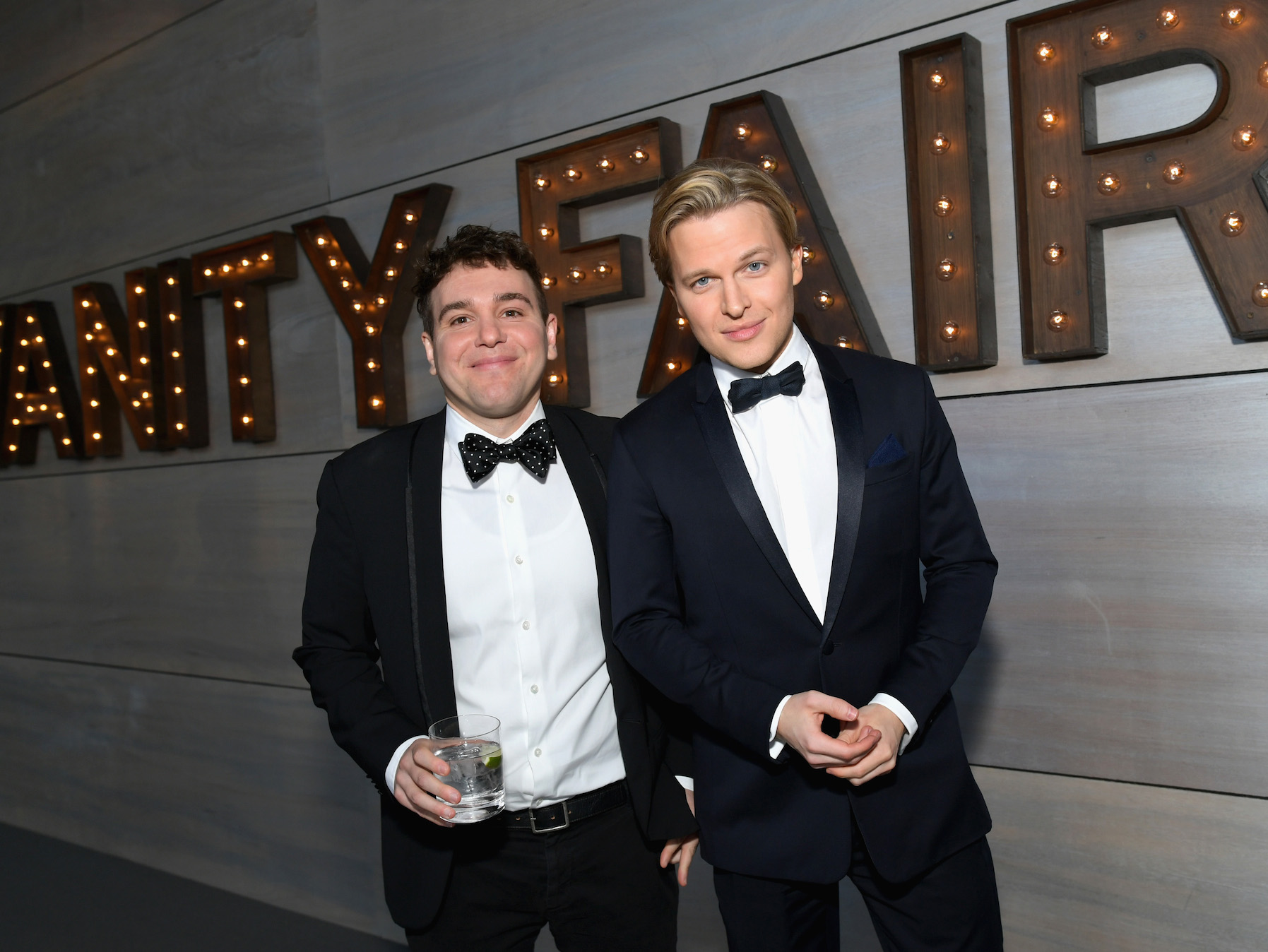 Jon Lovett and Ronan Farrow in suits standing in front of a Vanity Fair sign