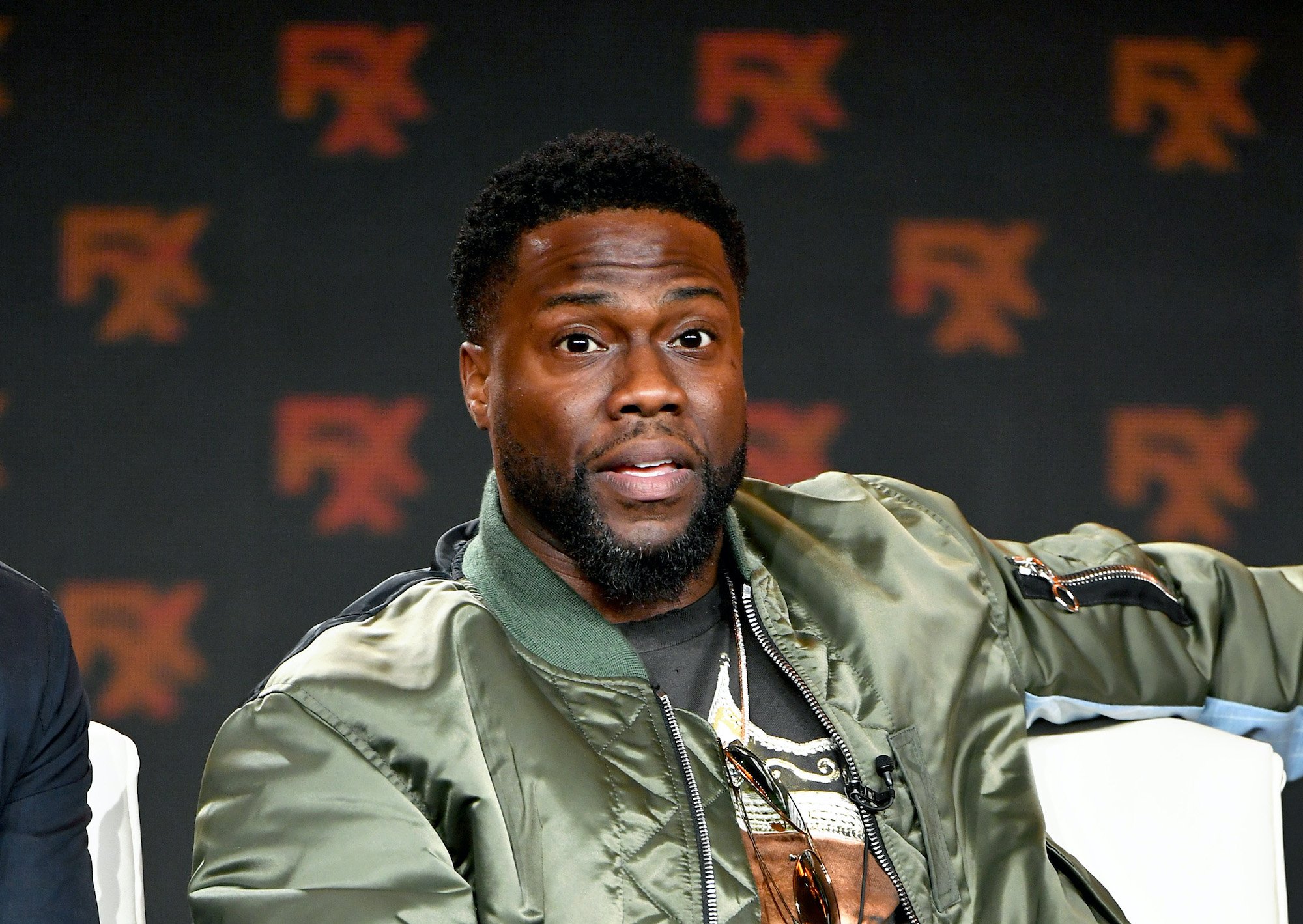Kevin Hart sitting in front of a black background with repeating logos