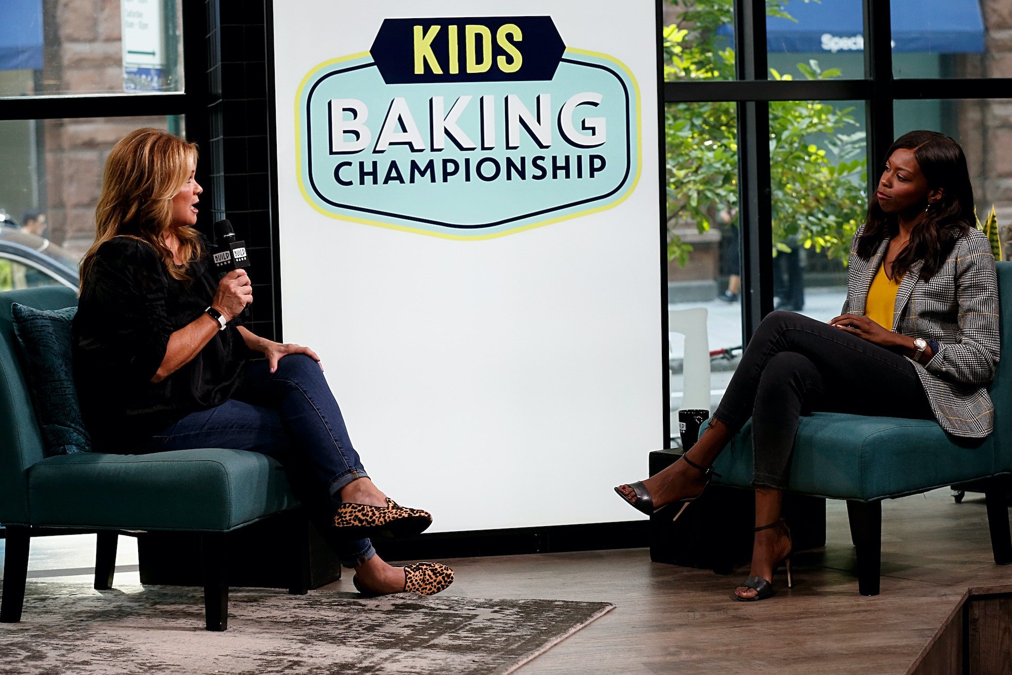 (L-R) Valerie Bertinelli and Brittany Jones-Cooper talking on a stage in front of a screen with the 'Kids Baking Championship' logo