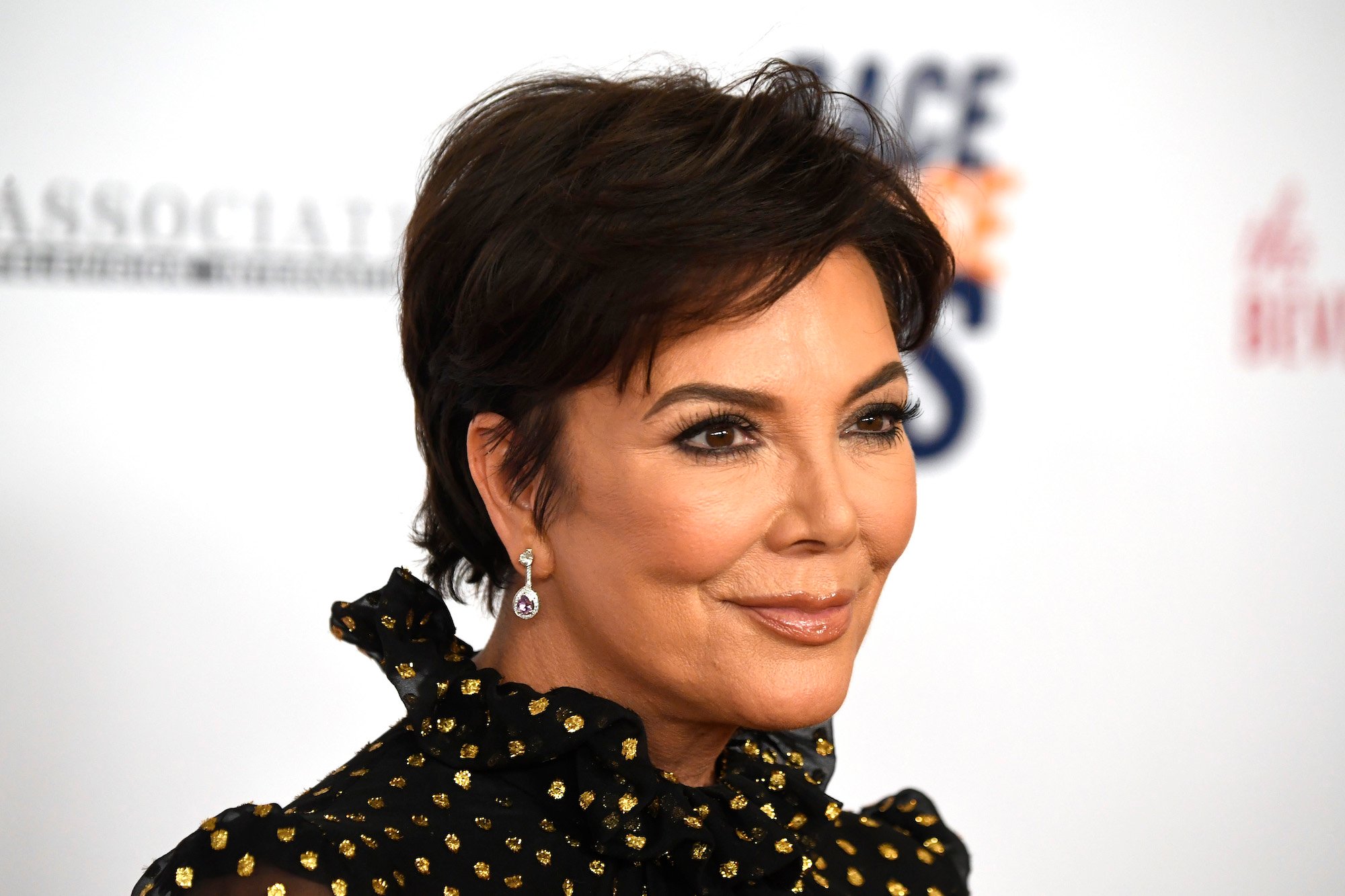 Kris Jenner smiling in front of a blurred white background, looking off camera