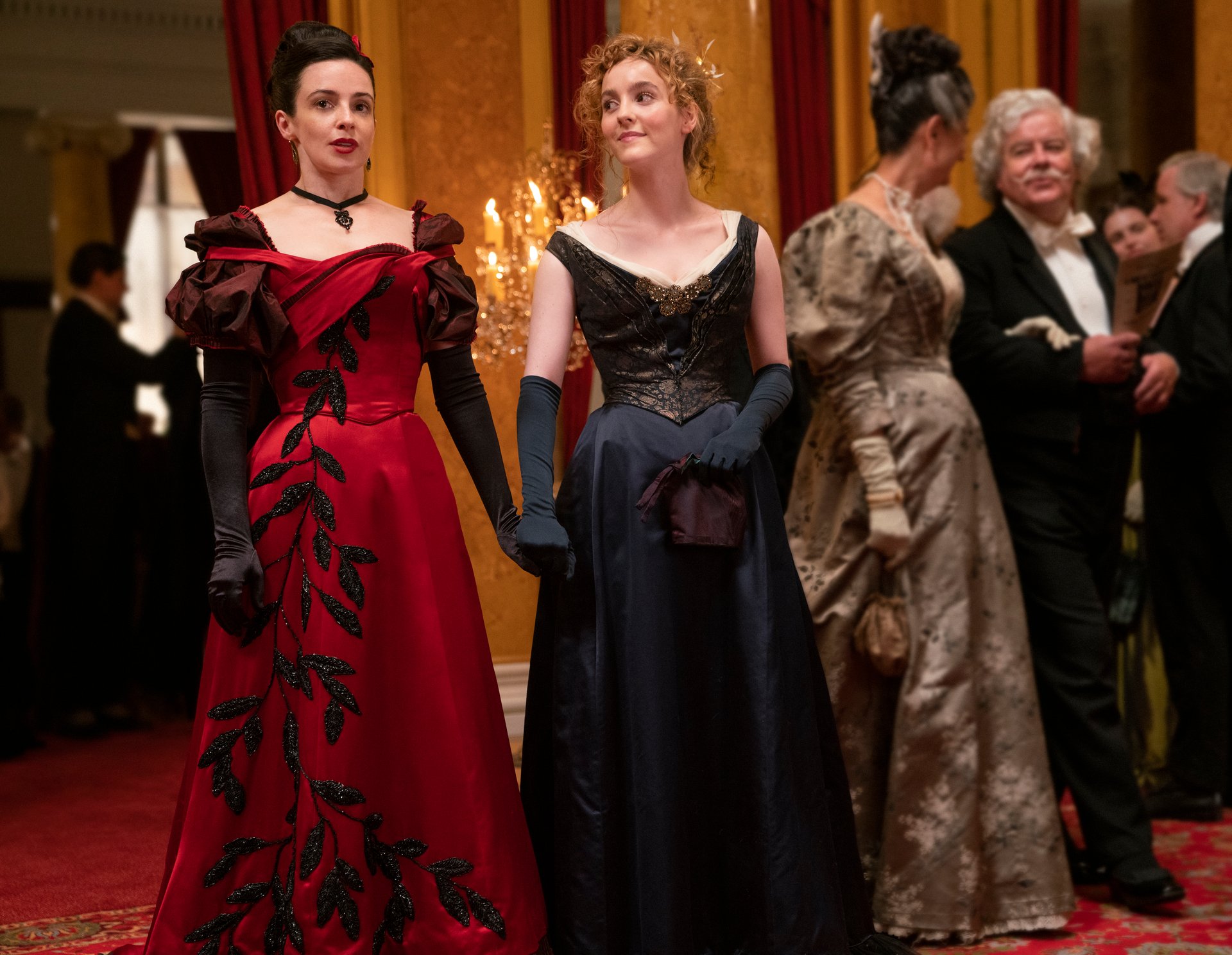 Laura Donnelly wearing a red dress holding hands with Ann Skelly wearing a black dress in The Nevers