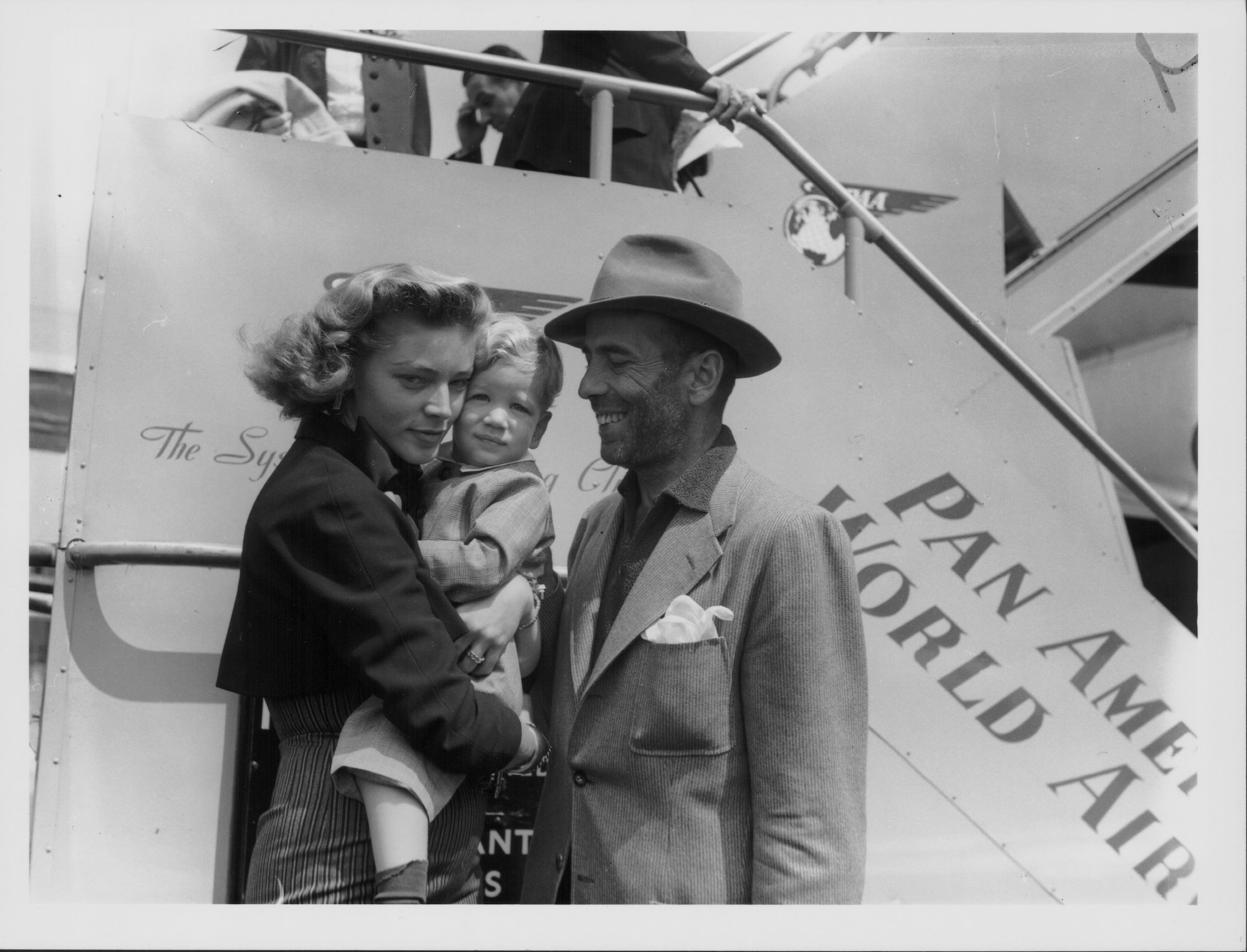 (L-R) Lauren Bacall holding a baby Stephen Humphrey Bogart, and Humphrey Bogart in front of a ship, smiling, in black and white