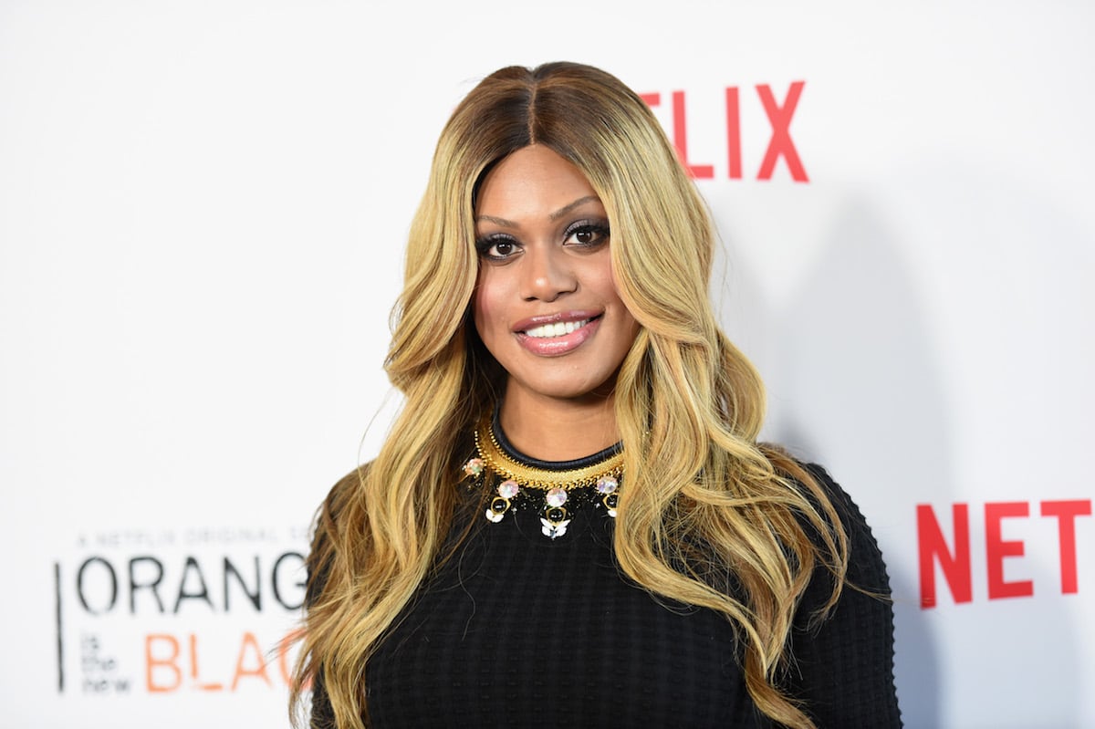 Laverne Cox staring at the camera while on the red carpet and smiling. She's wearing black and has her blonde hair down.