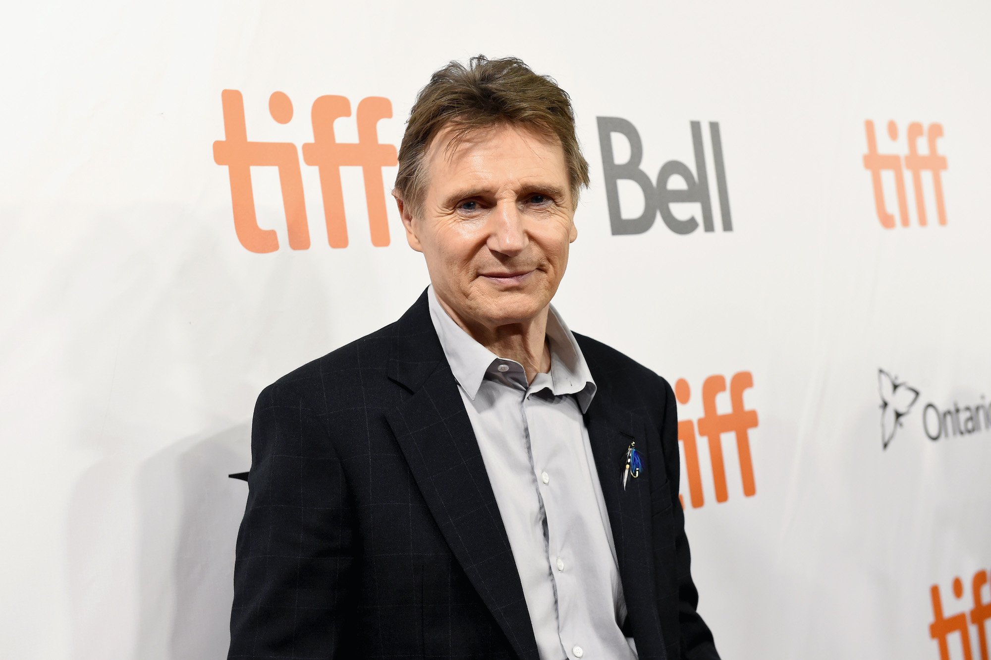 Liam Neeson smiling in front of a white background with repeating logos