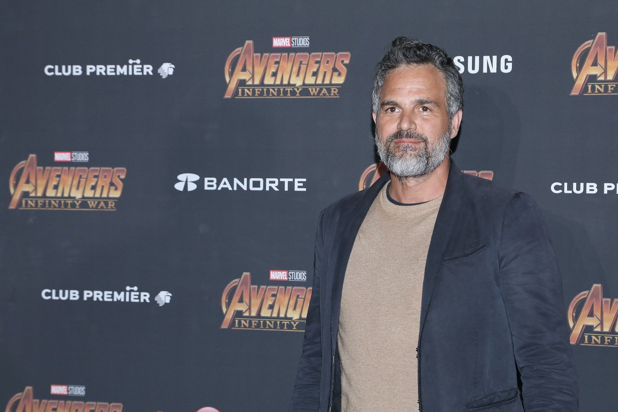 Mark Ruffalo smiling in front of a gray background with repeating logos