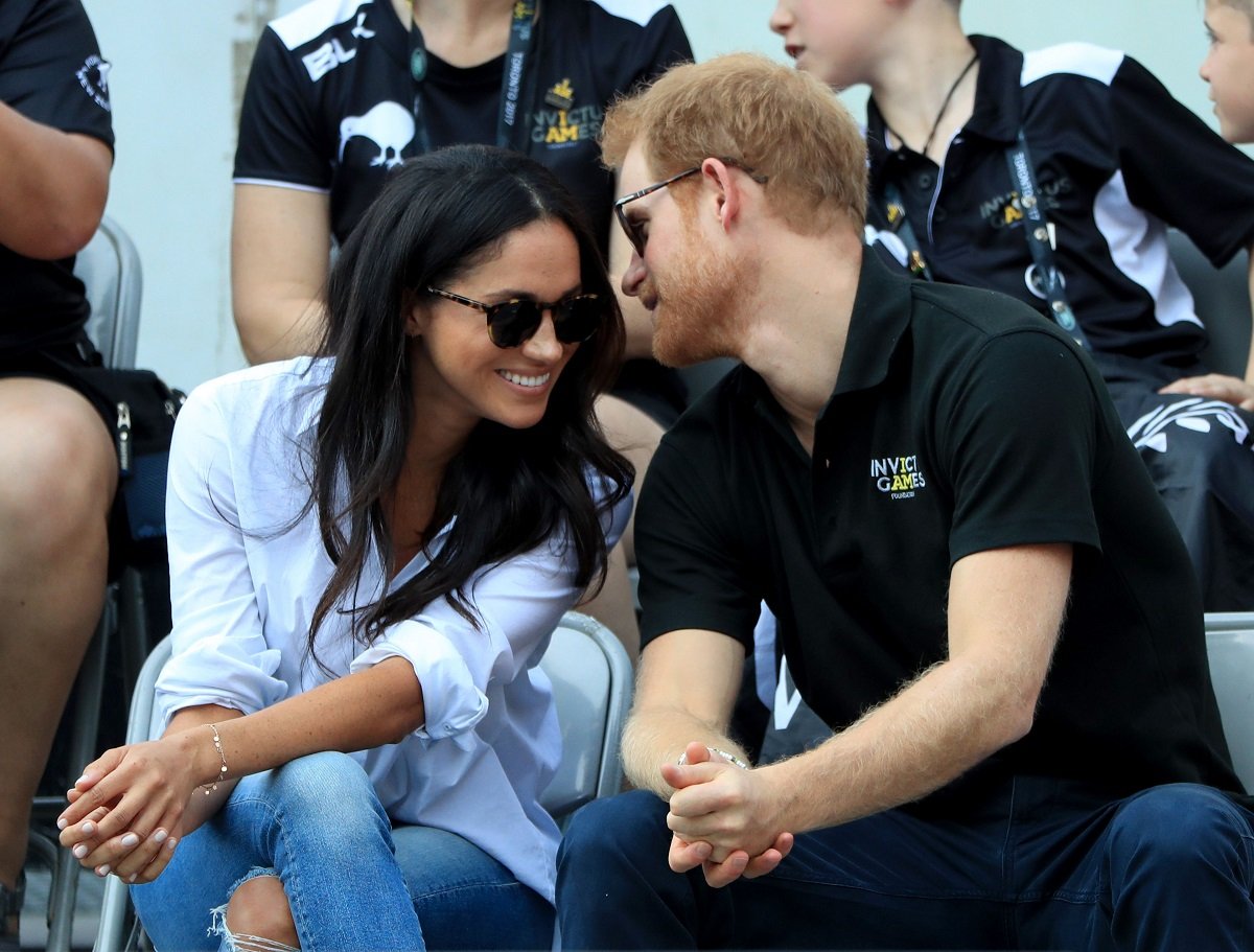 Prince Harry and Meghan Markle wearing sunglasses and leaning in to each other while watching Wheelchair Tennis in 2017