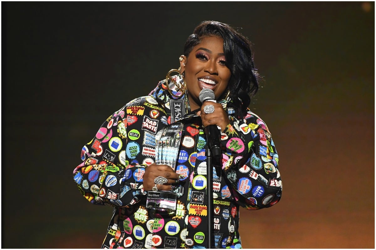 Missy Elliott smiling at an awards show wearing a rainbow jacket and gold earrings. 