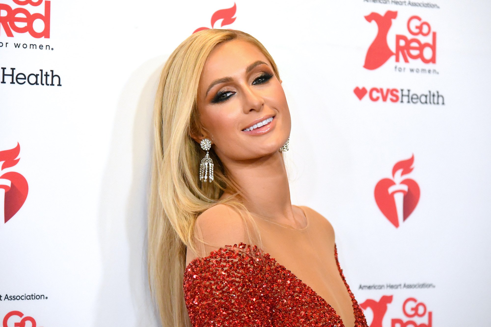 Paris Hilton smiling in front of a white background with repeating red logos