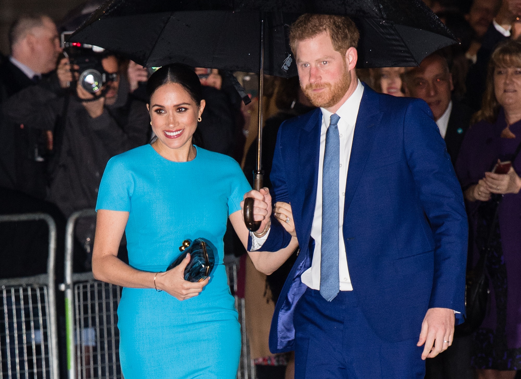 Prince Harry and Meghan Markle stand under an umbrella with Meghan wearing a turquoise dress and Harry in a blue suit