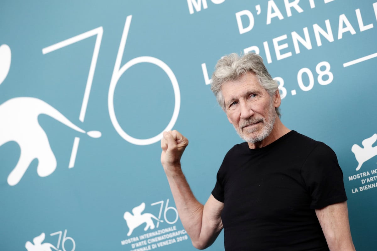 Roger Waters photo of his upper body wearing a black t-shirt and holding up his right hand.