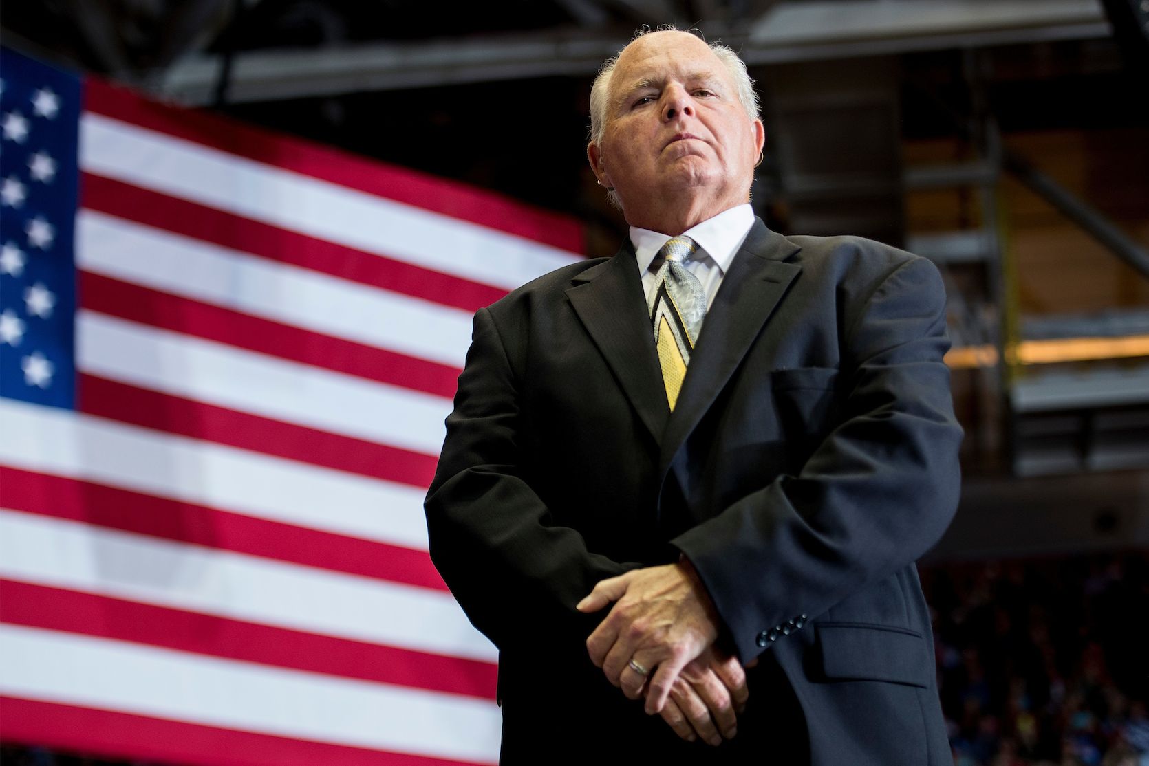 Rush Limbaugh standing in front of an American flag with a serious look on his face