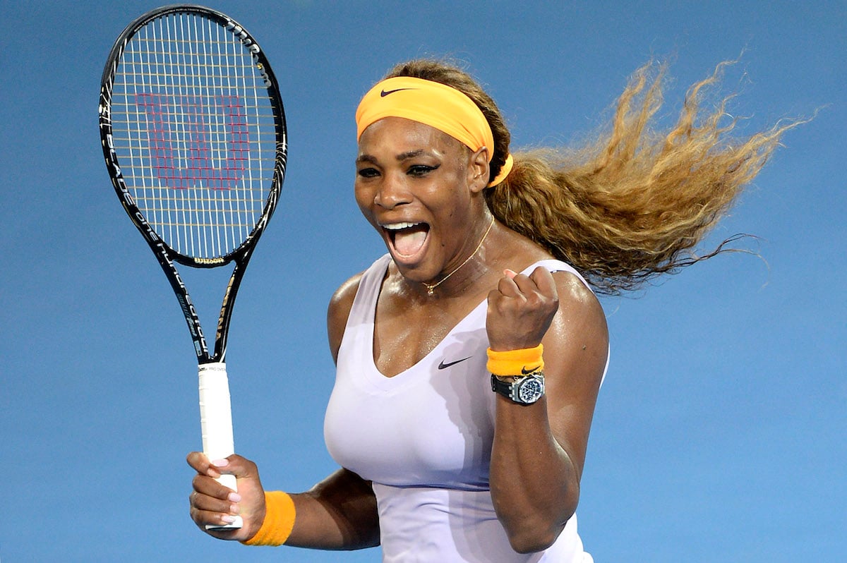 Serena Williams on the tennis court wearing white, a yellow headband and holding a tennis racket in her right hand.