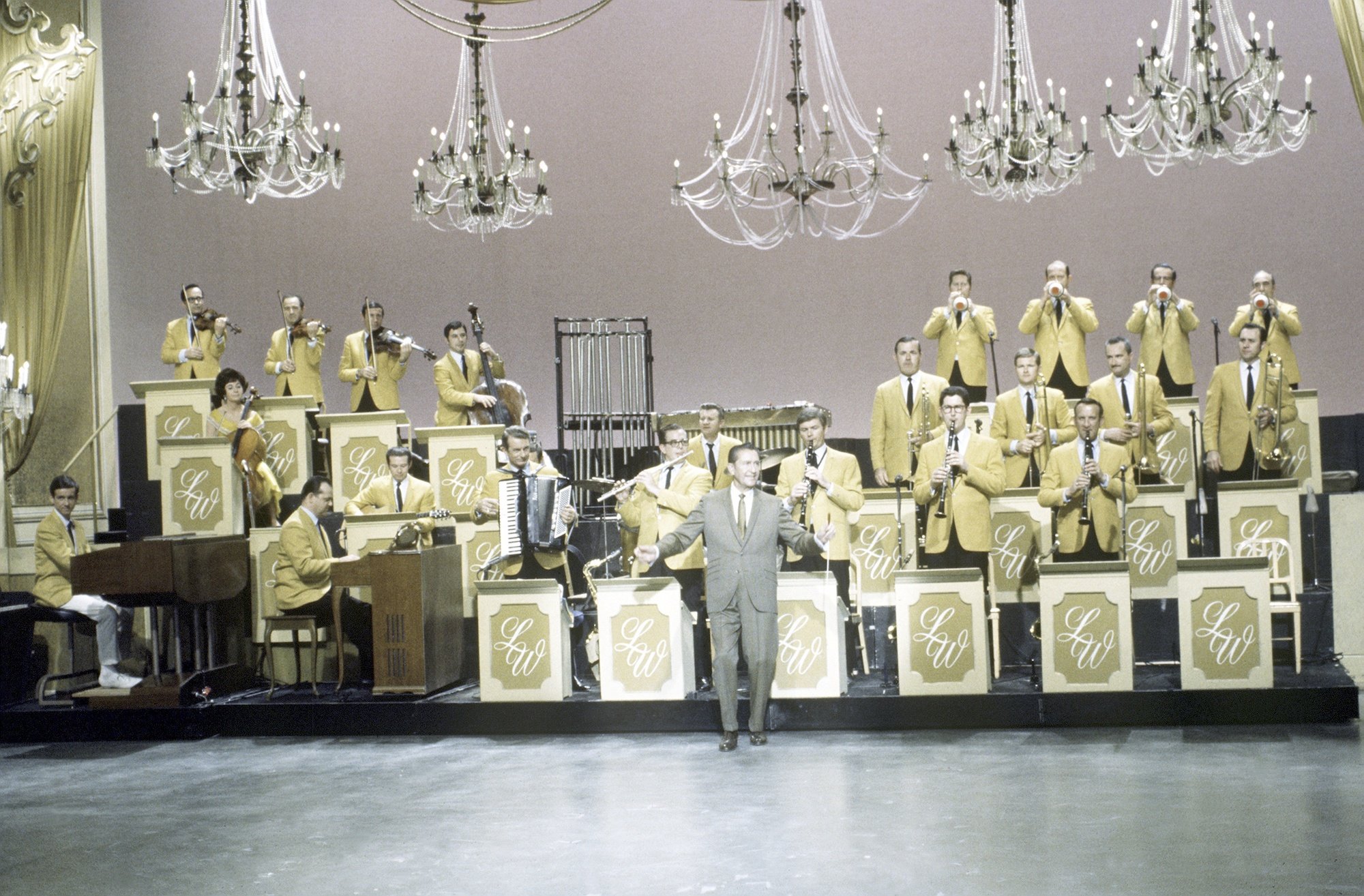 Lawrence Welk on stage in front of a band, under chandeliers