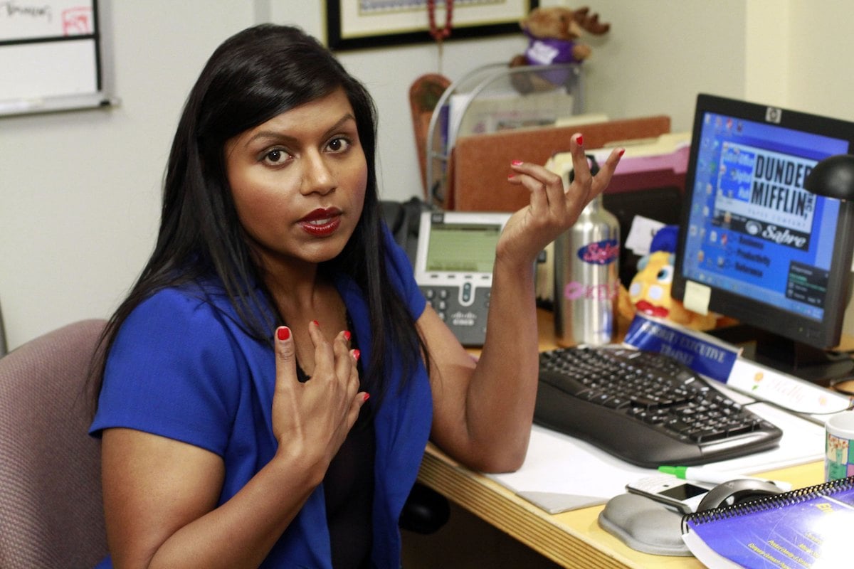 Mindy Kaling as Kelly Kapoor looks at the camera siting at a desk with her hands up on 'The Office'