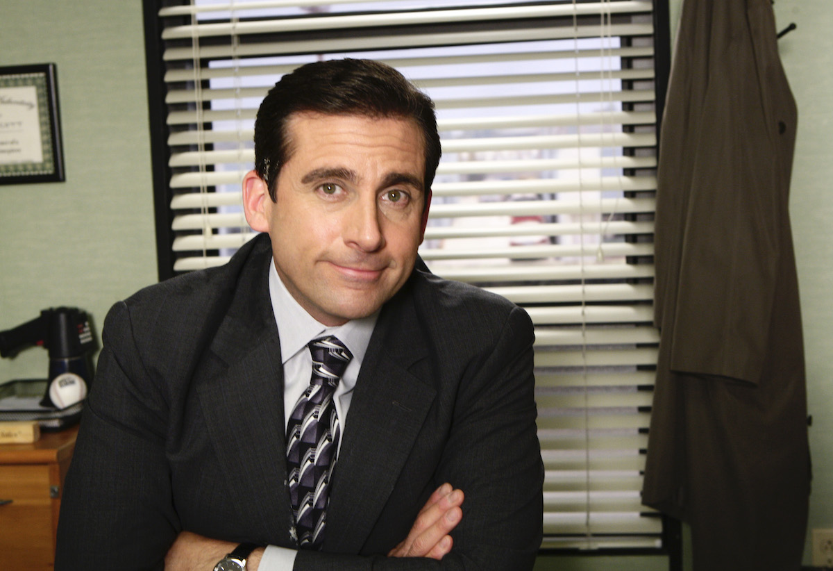 Steve Carell as Michael Scott smiles with his arms crossed in front of him as he sits in front of a window on 'The Office'