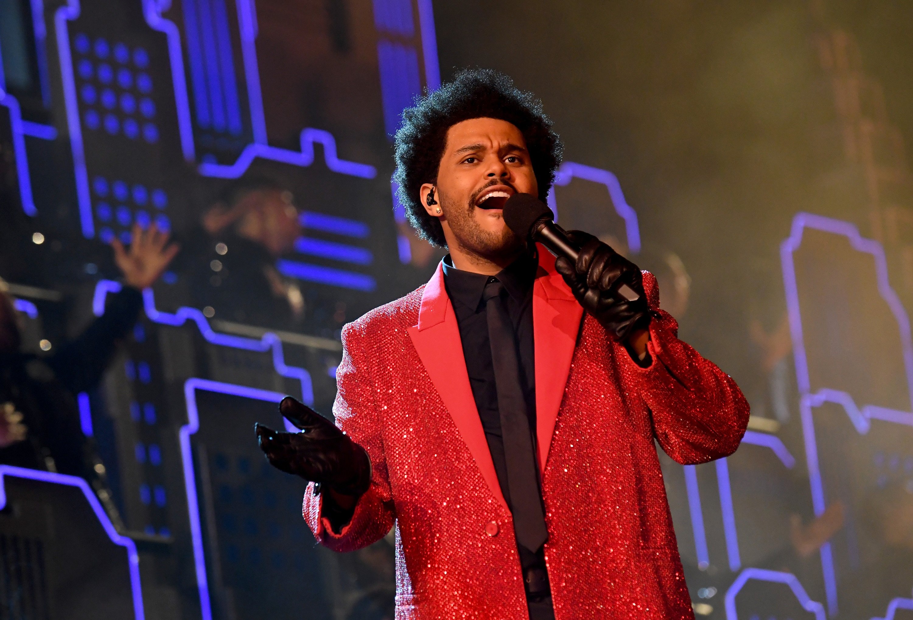 The Weeknd holds a microphone singing in a red jacket and black gloves at the Super Bowl halftime show