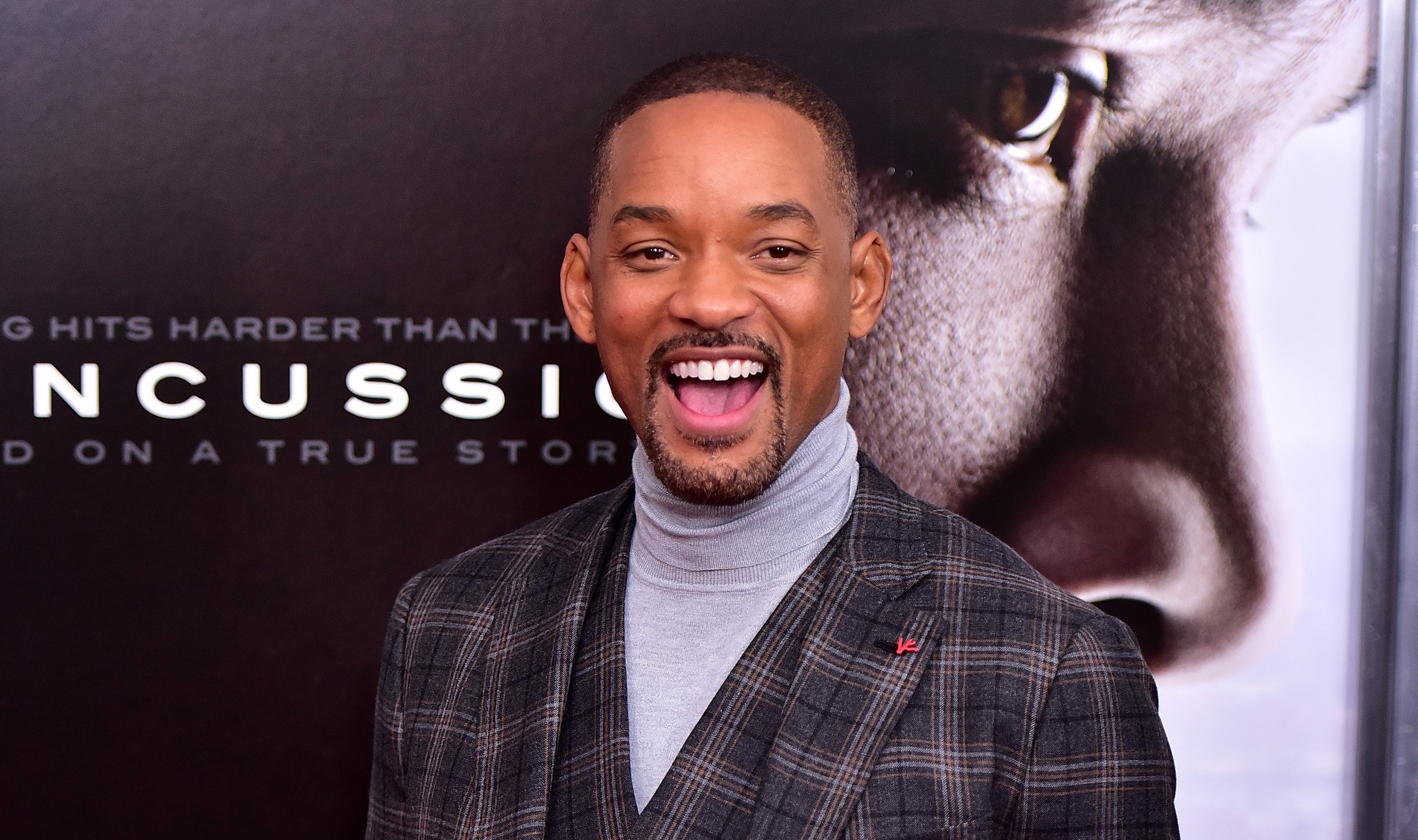 Will Smith smiling at a movie premiere wearing a grey turtleneck and plaid blazer.