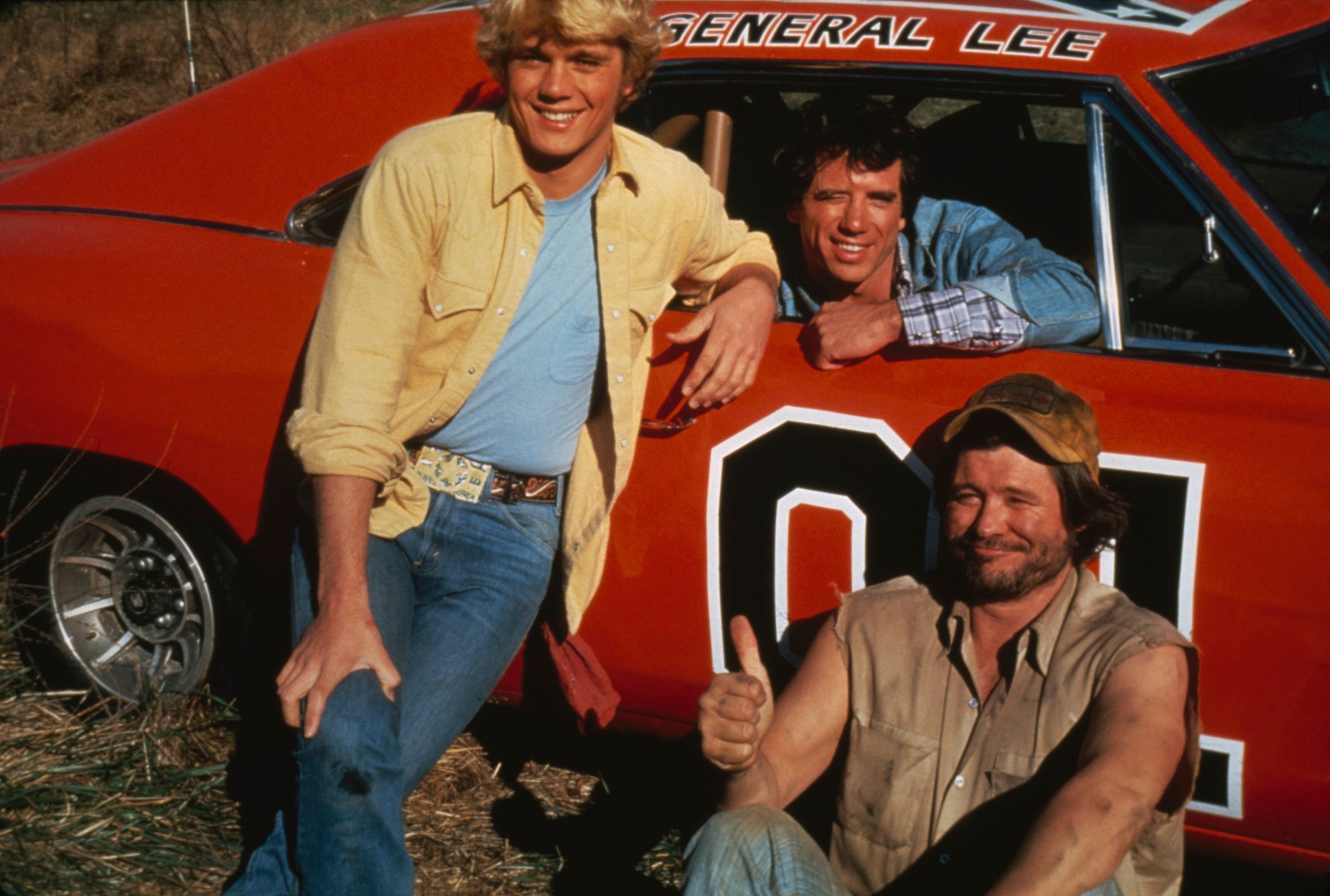 John Schneider, Tom Wopat, and Ben Jones as Bo Duke, Luke Duke, and Cooter. They are posing with the General Lee in The Dukes of Hazzard