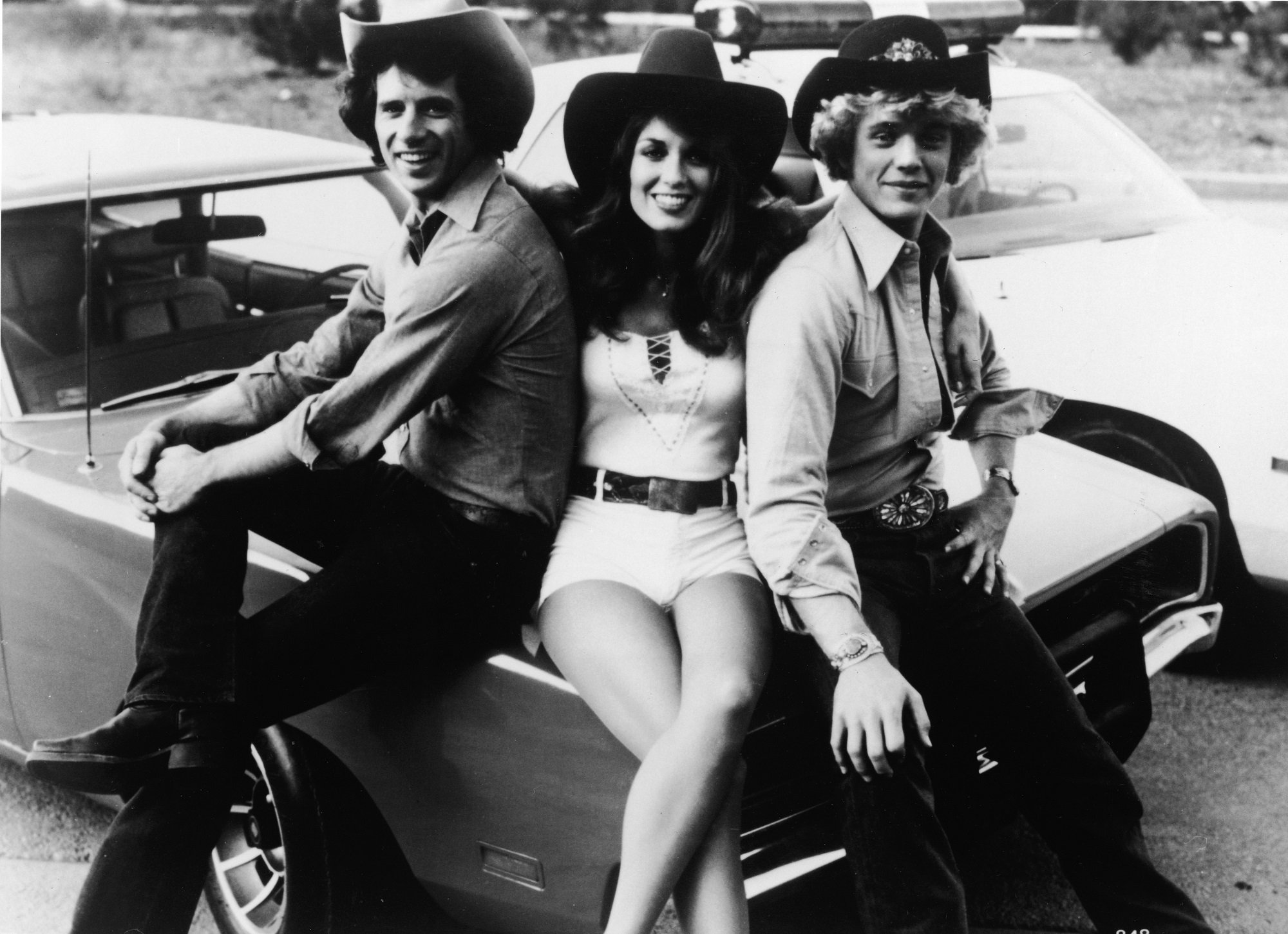 Tom Wopat, Catherine Bach, and John Schneider sitting on the hood of the car, the General Lee for the television show, The Dukes of Hazzard