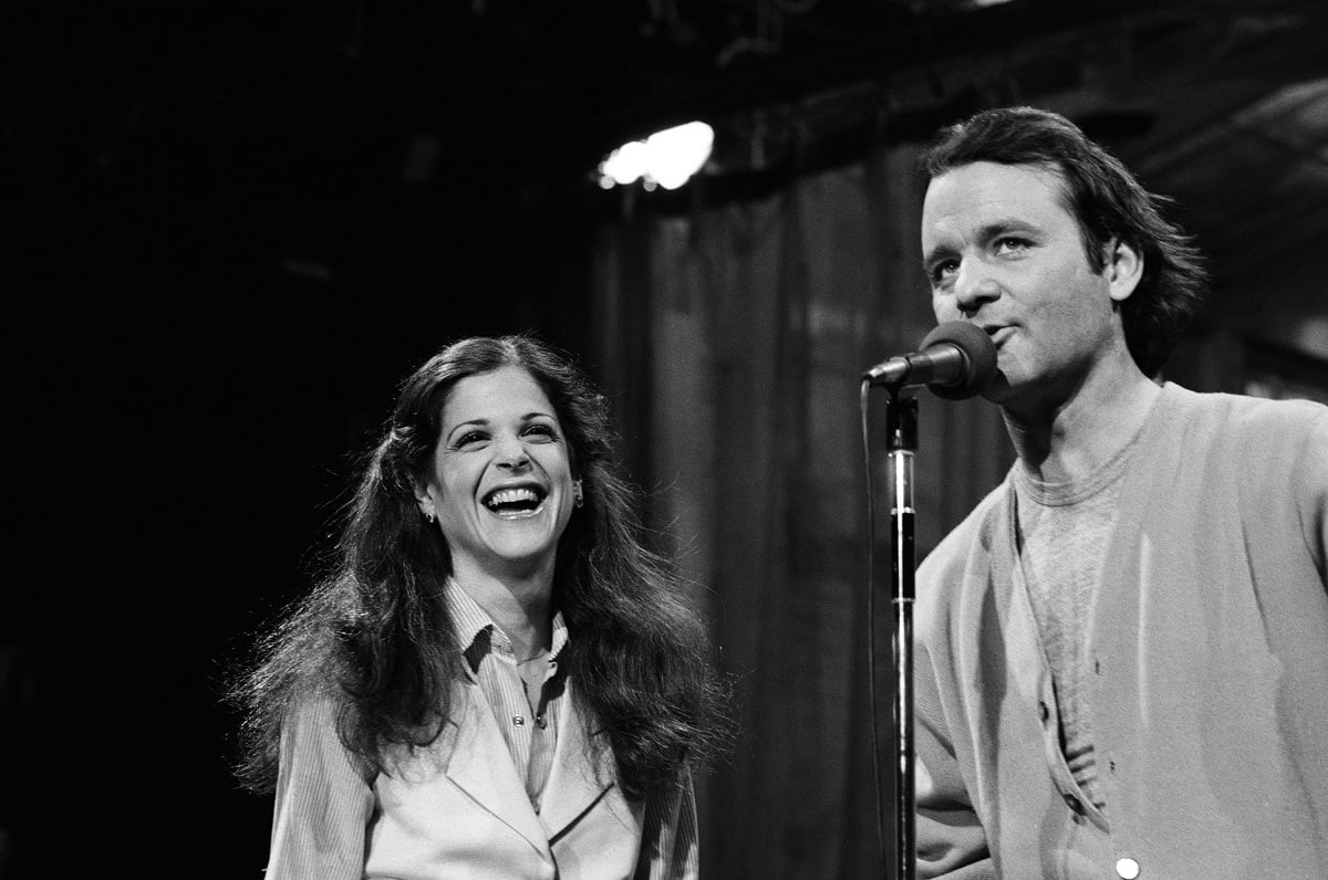 A black-and-white photo of Gilda Radner laughing at Bill Murray on the mic on 'Saturday Night Live'
