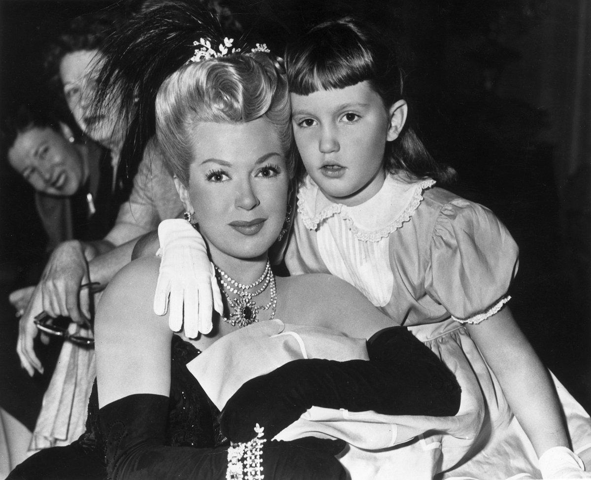 Lana Turner (L), wearing a period costume with a feather headdress, sits with her daughter, Cheryl Crane, on the set of 'The Merry Widow' in 1952.