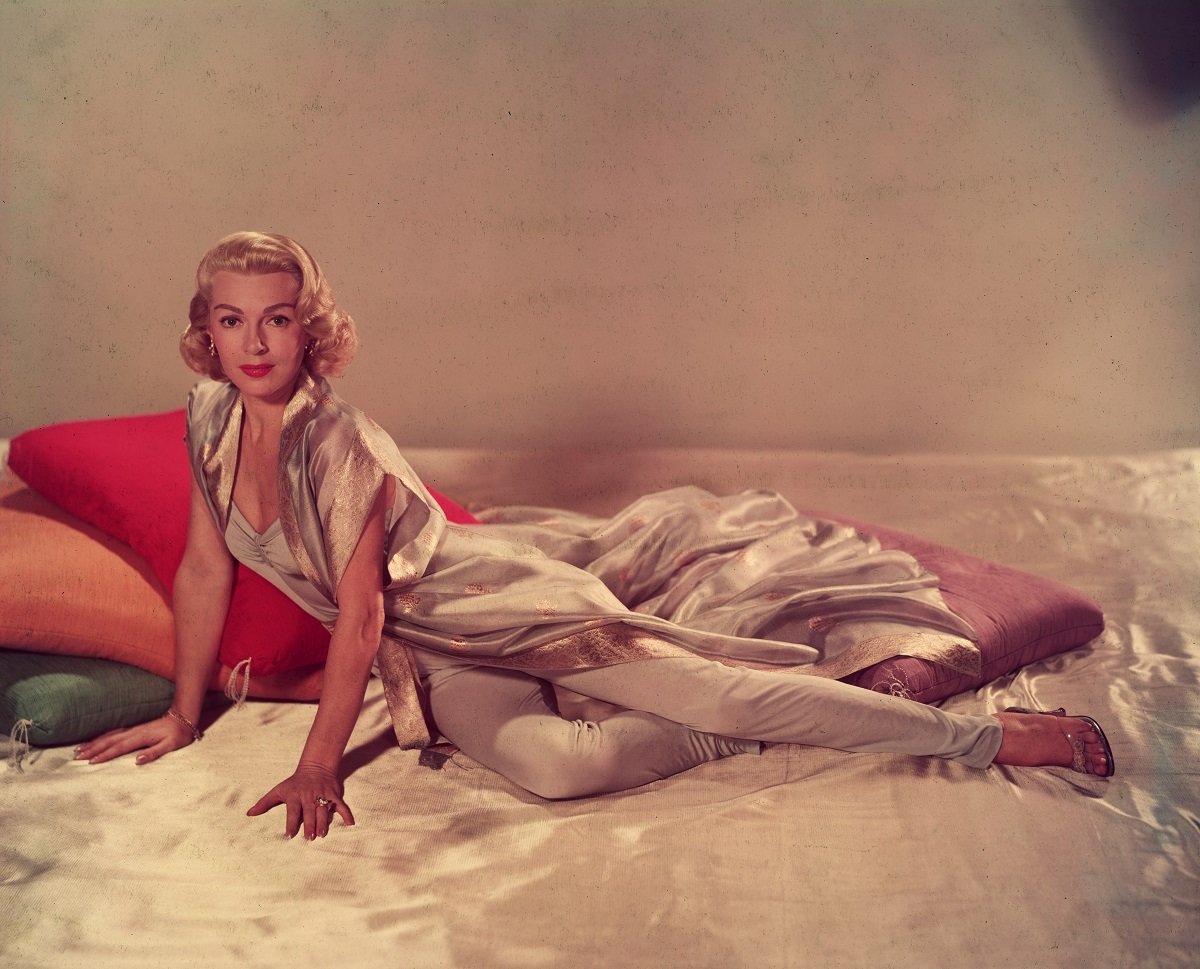 Lana Turner in 1955 posing on a silk sheet with large cushions in a sleeveless v-neck jumpsuit and a short-sleeved silk robe.
