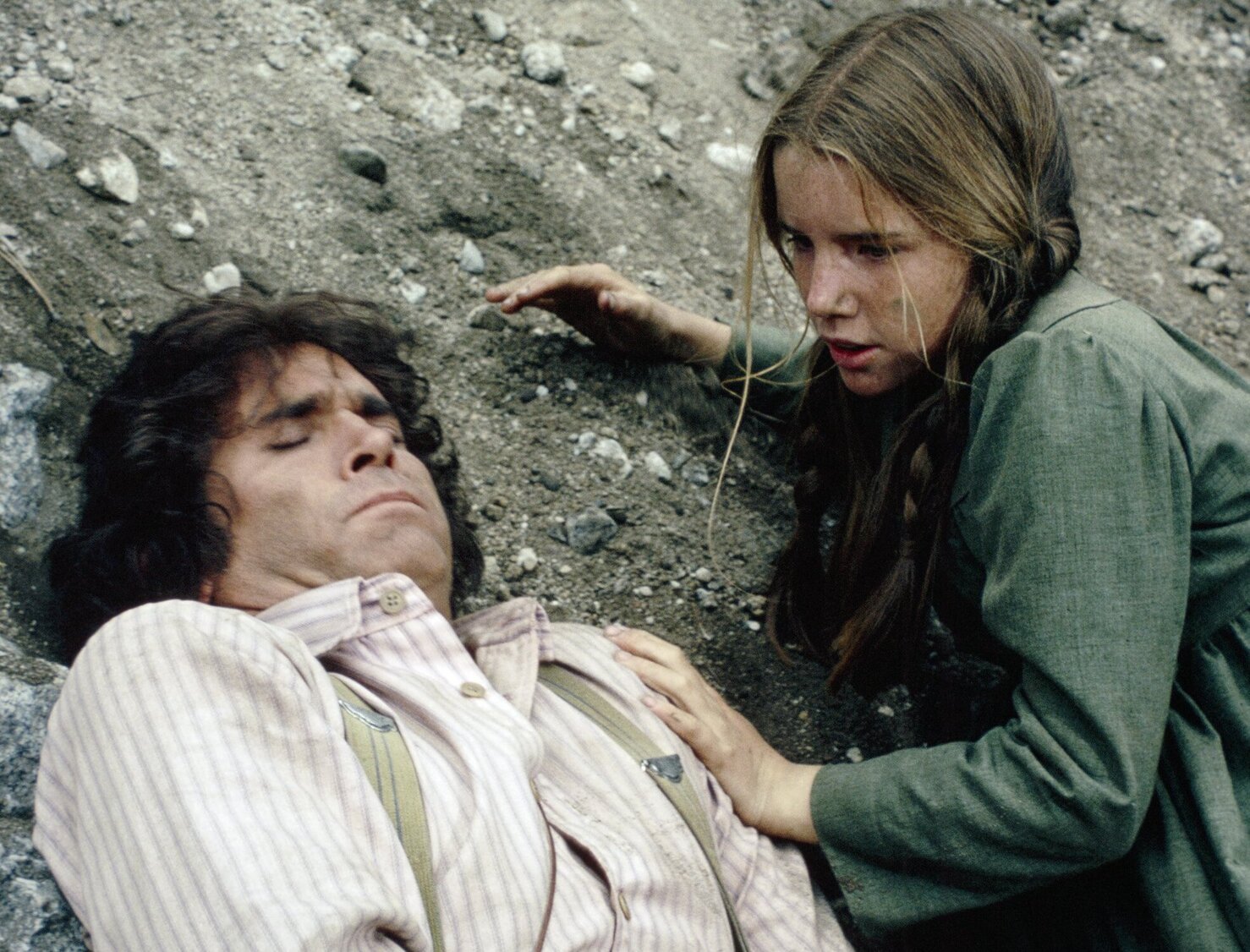 Michael Landon as Charles Ingalls lying in the dirt as Melissa Gilbert as Laura Ingalls bends down beside him.