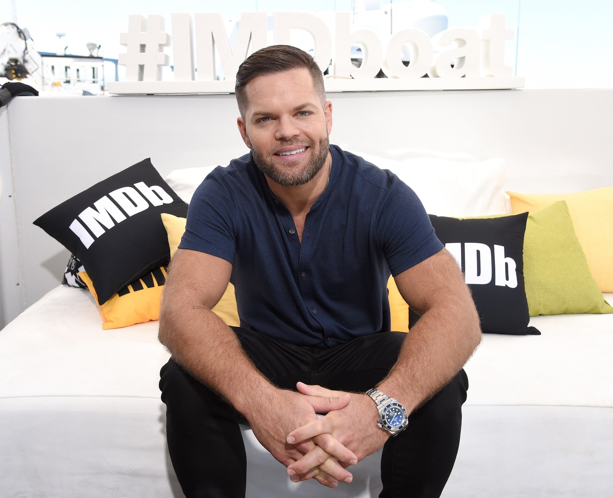 The Expanse star Wes Chatham sits on a white coach with IMDb throw pillows