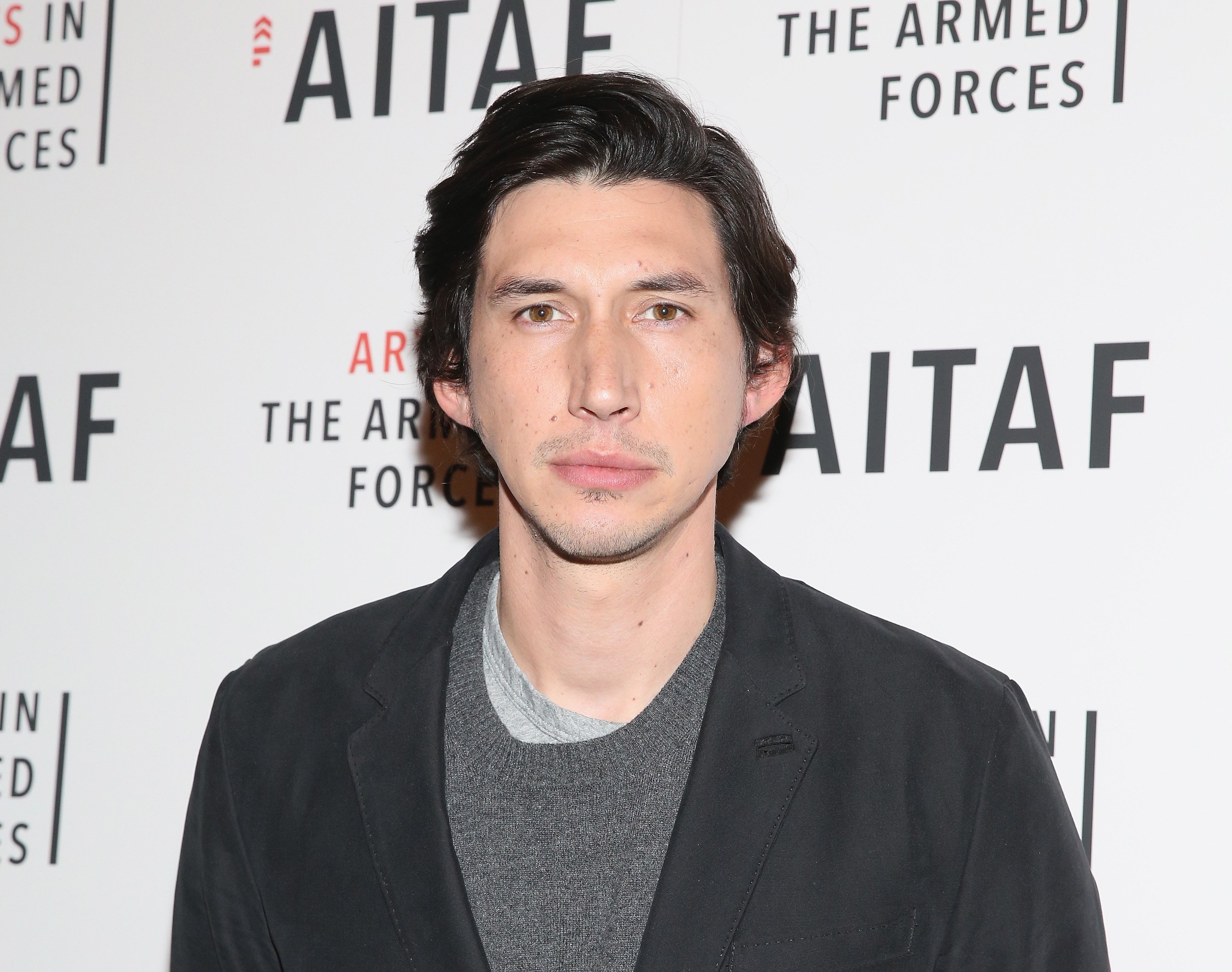 Adam Driver at a photo opportunity. He's looking at the camera, not smiling, wearing a grey sweater and black blazer.