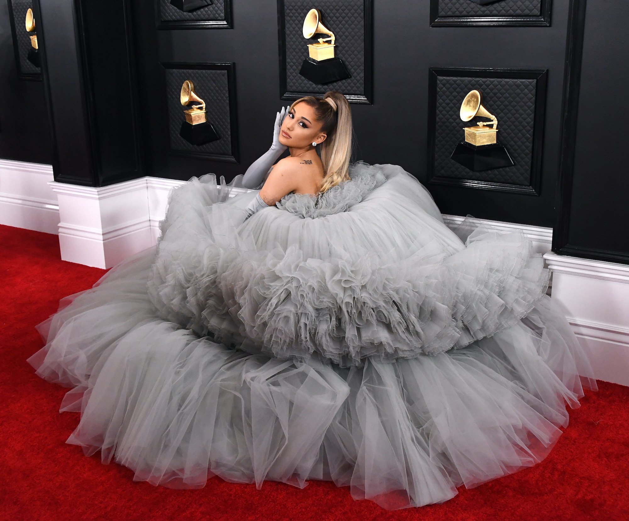 Ariana Grande crouched on a red carpet in a gray dress