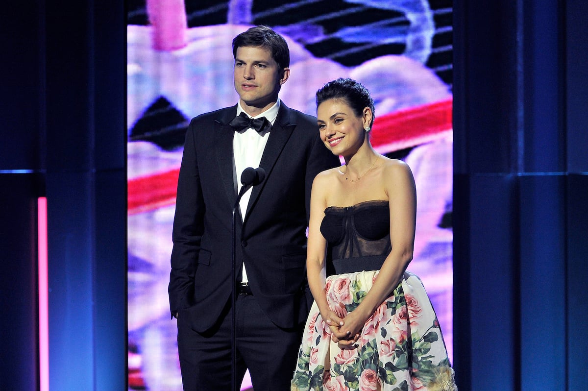 Ashton Kutcher and Mila Kunis stand next to each other onstage in front of a mic at the 2018 Breakthrough Prize