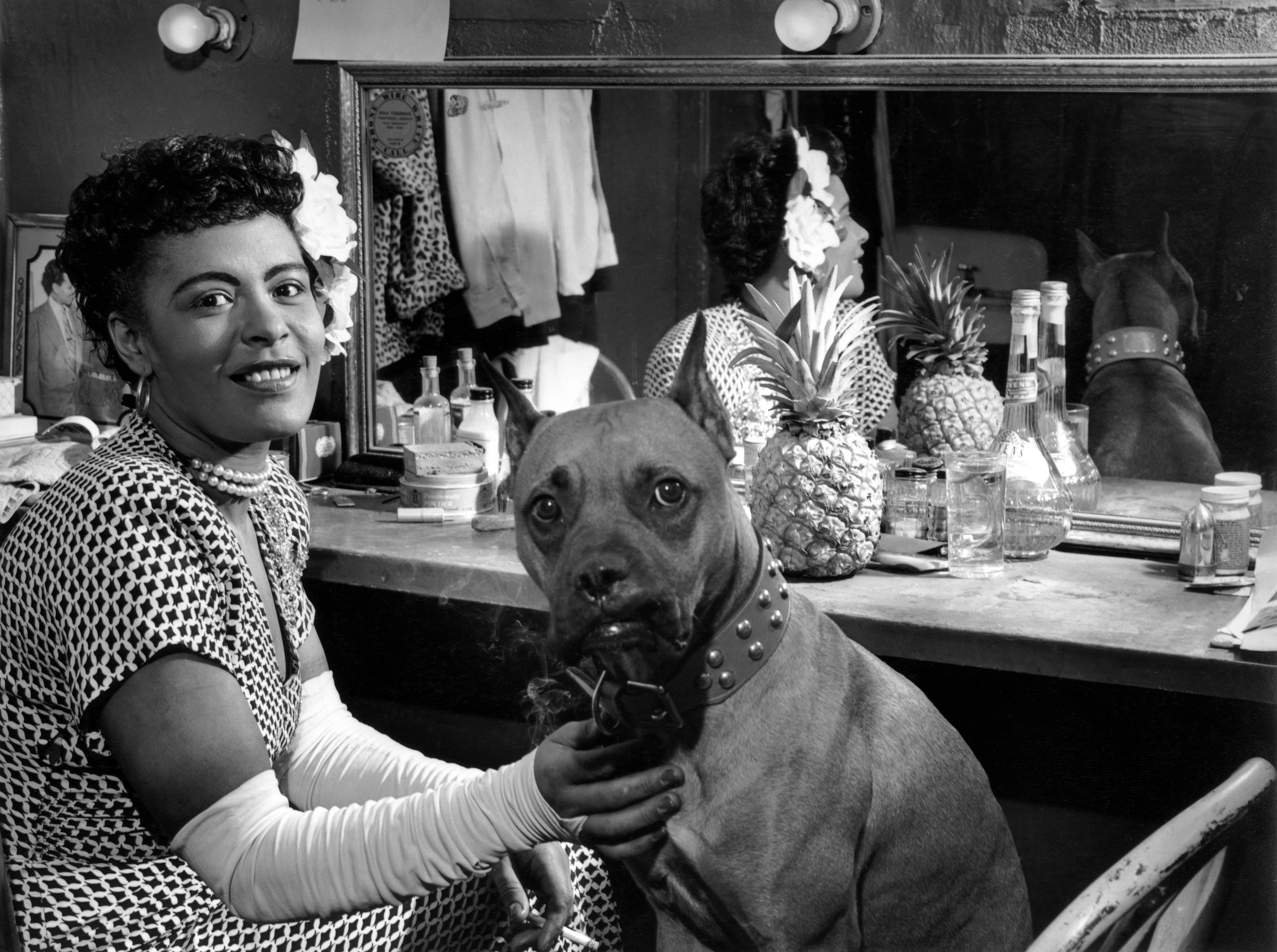 A black and white photo of Billie Holiday smiling backstage with her dog.
