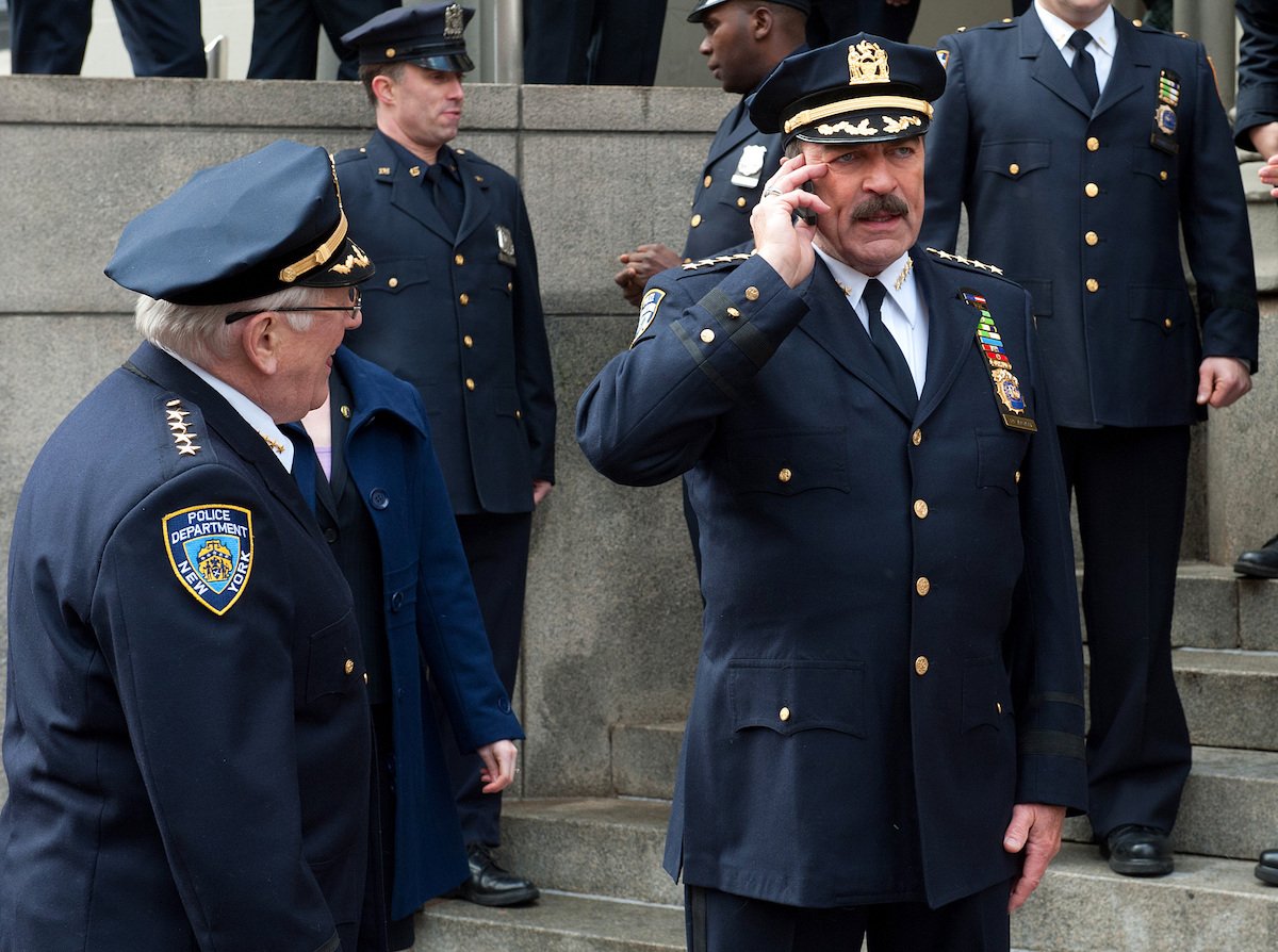 Len Cariou as Henry Reagan and Tom Selleck as Frank Reagan on 'Blue Bloods' stand outside in their police uniforms. Frank is on the phone.