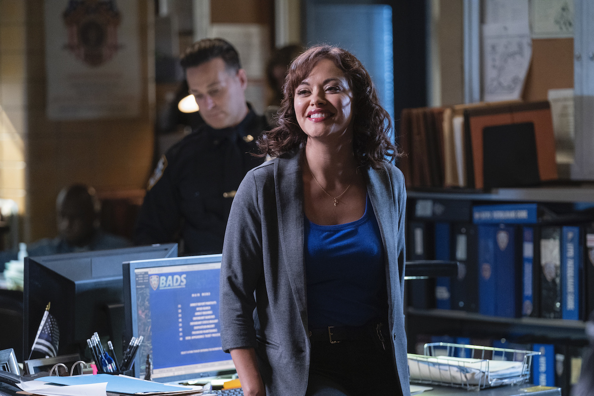 Marisa Ramirez as Maria Baez leans on a desk in a police precinct smiling on 'Blue Bloods'