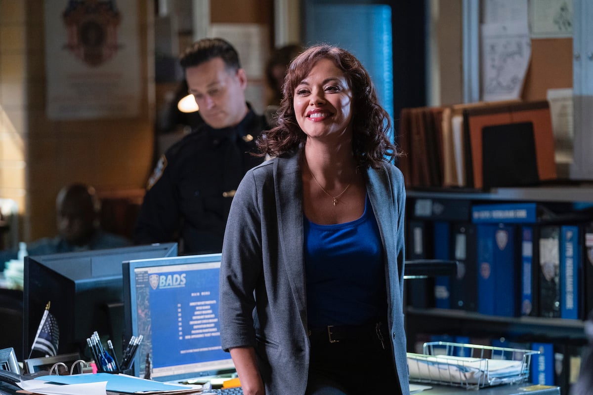 Marisa Ramirez as Maria Baez leans on a desk in a police precinct smiling on 'Blue Bloods'