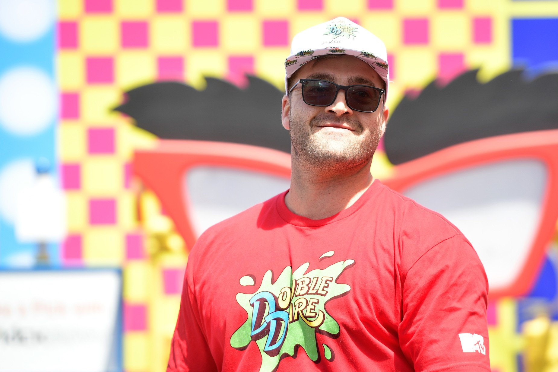 CT Tamburello from MTV's 'The Challenge' wearing a hat and sunglasses along with a 'Double Dare' shirt smiling 