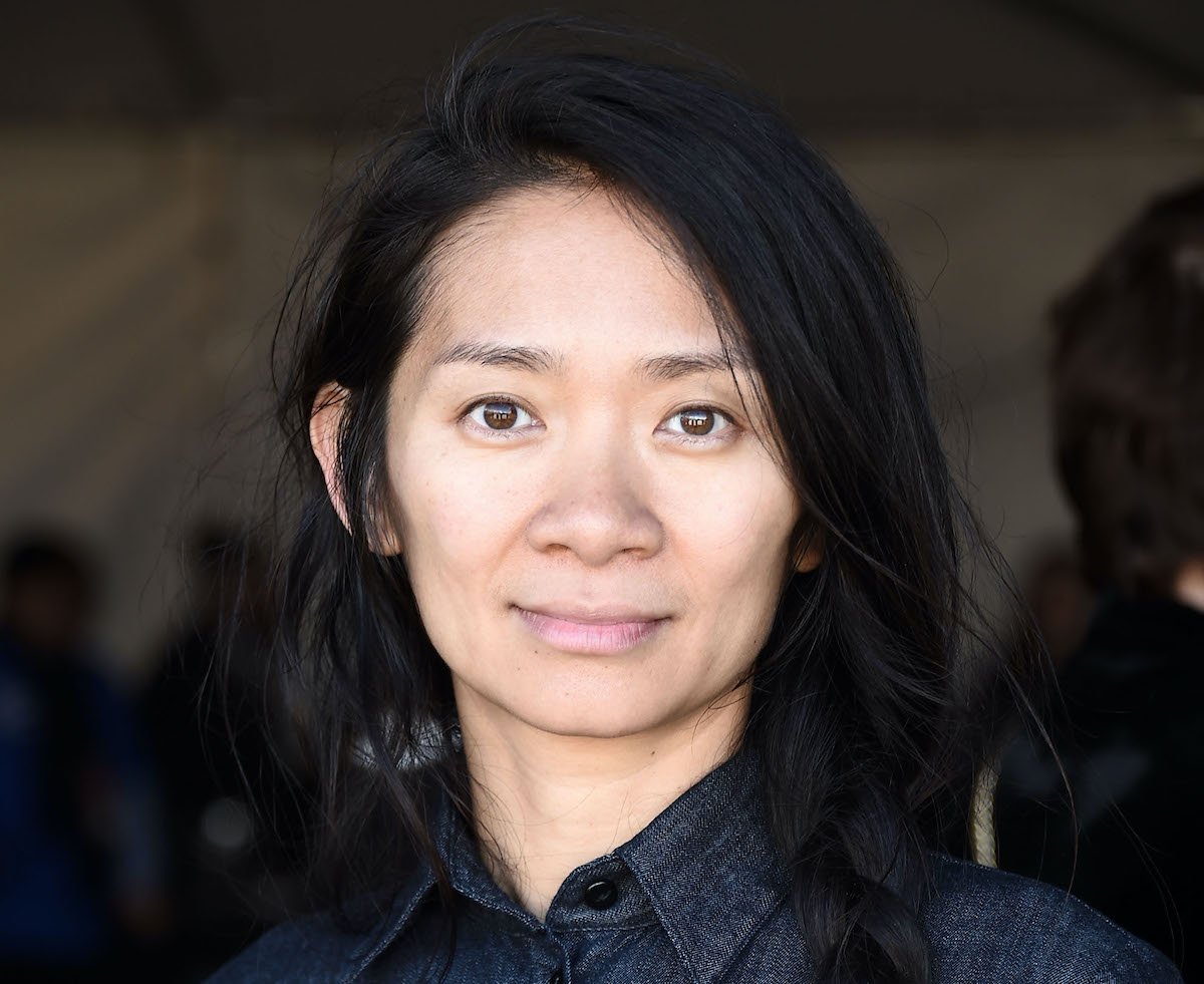 Chloé Zhao poses in a dark denim dress with her long black hair in a braid | Amanda Edwards/Getty Images