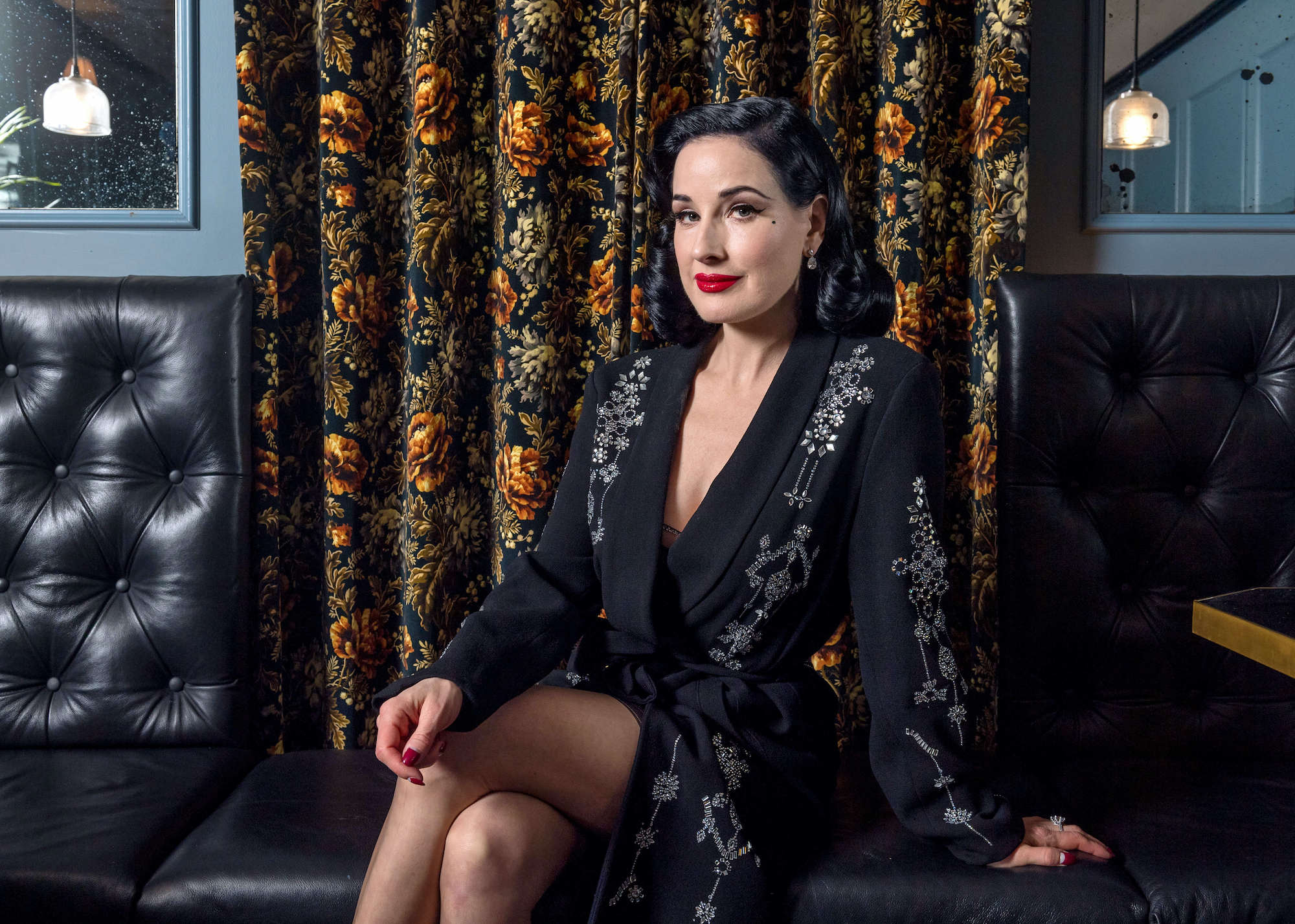 Dita Von Teese sitting on a couch in front of a floral curtain