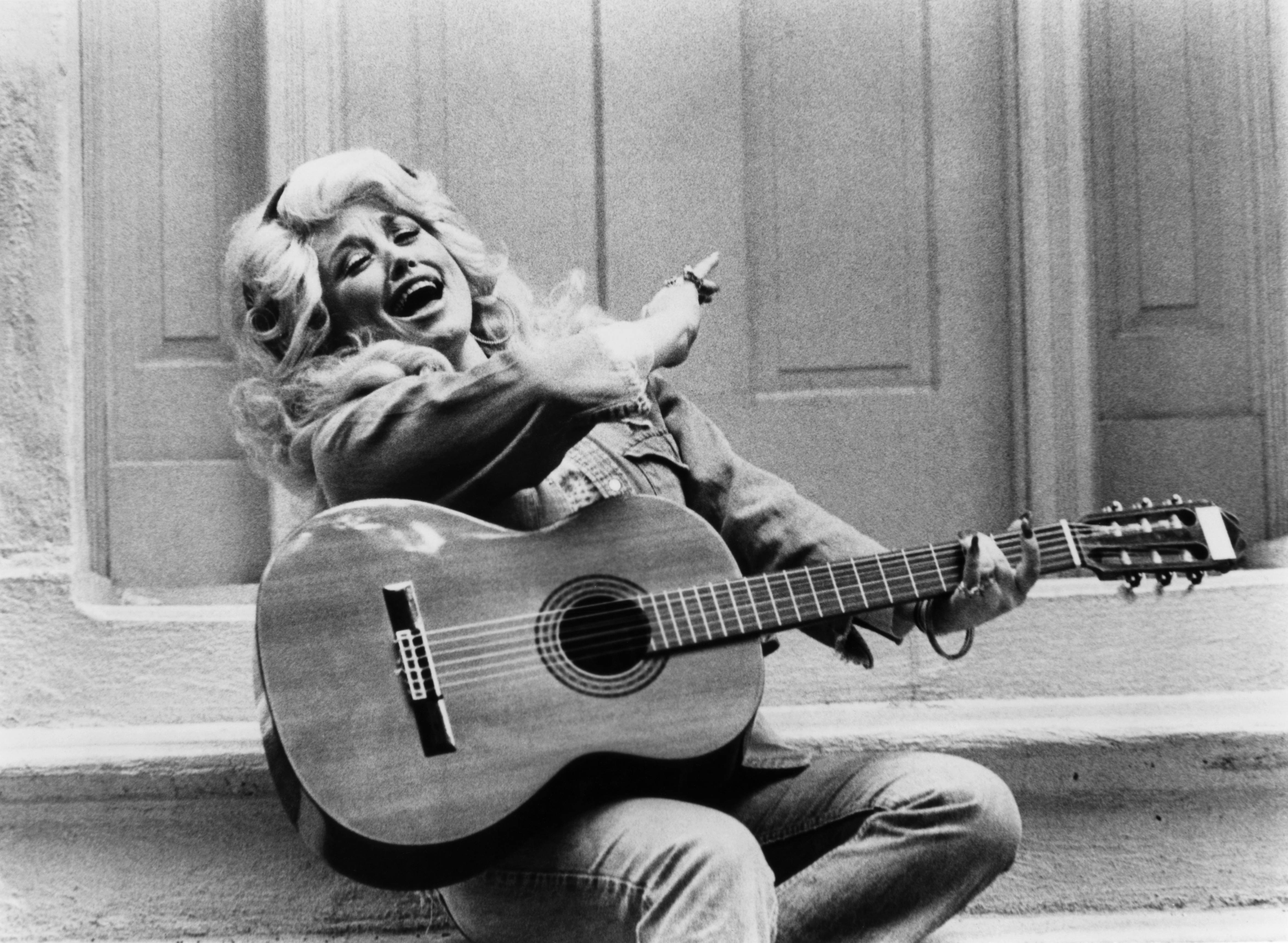 Dolly Parton spiritedly plays the guitar in black and white in 1970. She's wearing her hair big and curl and a jean outfit.