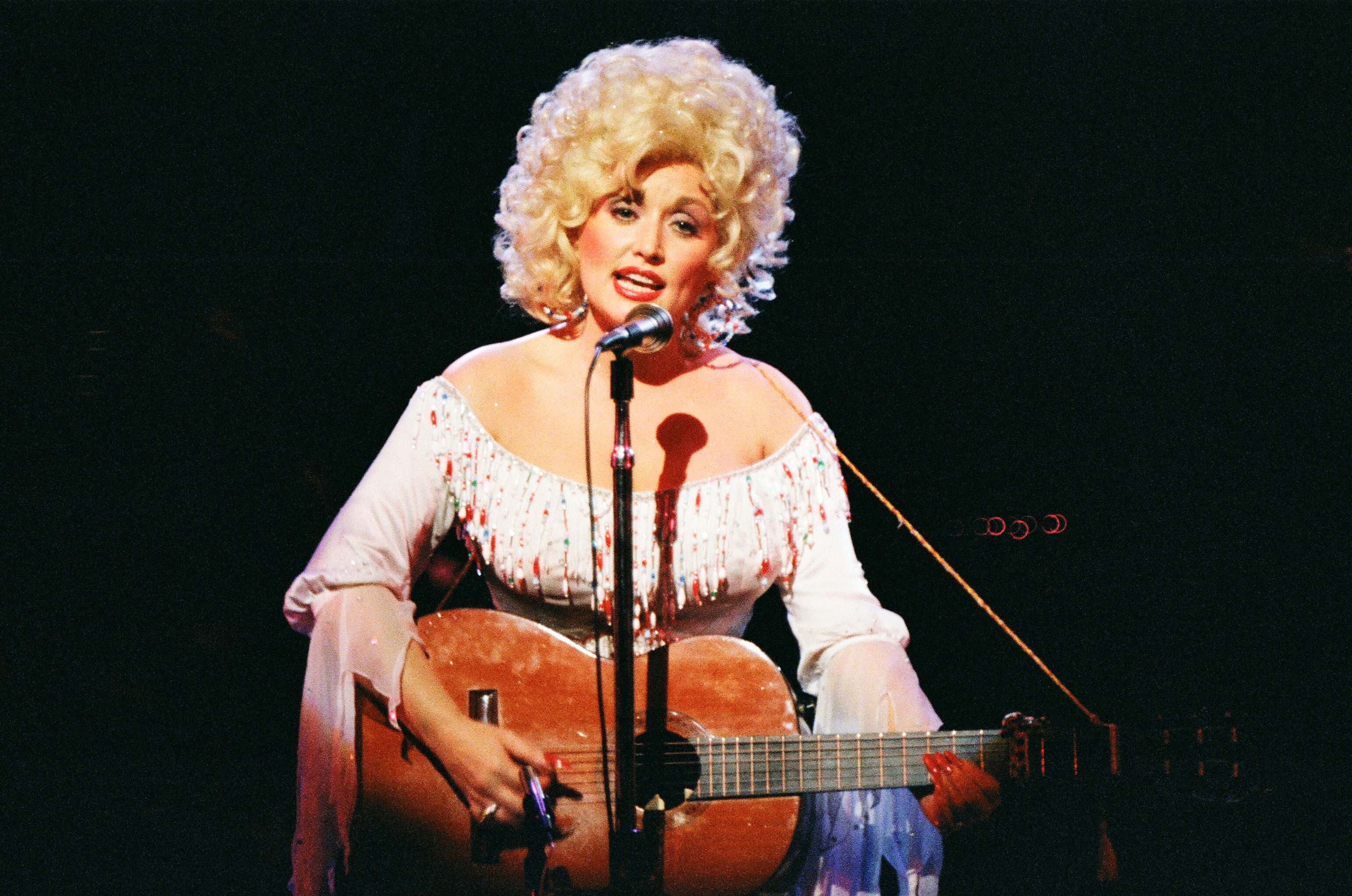 Dolly Parton performs at the Dominion Theatre in London in a sparkly white top and big, blond, curly hair. She's strumming a guitar and singing into a microphone.