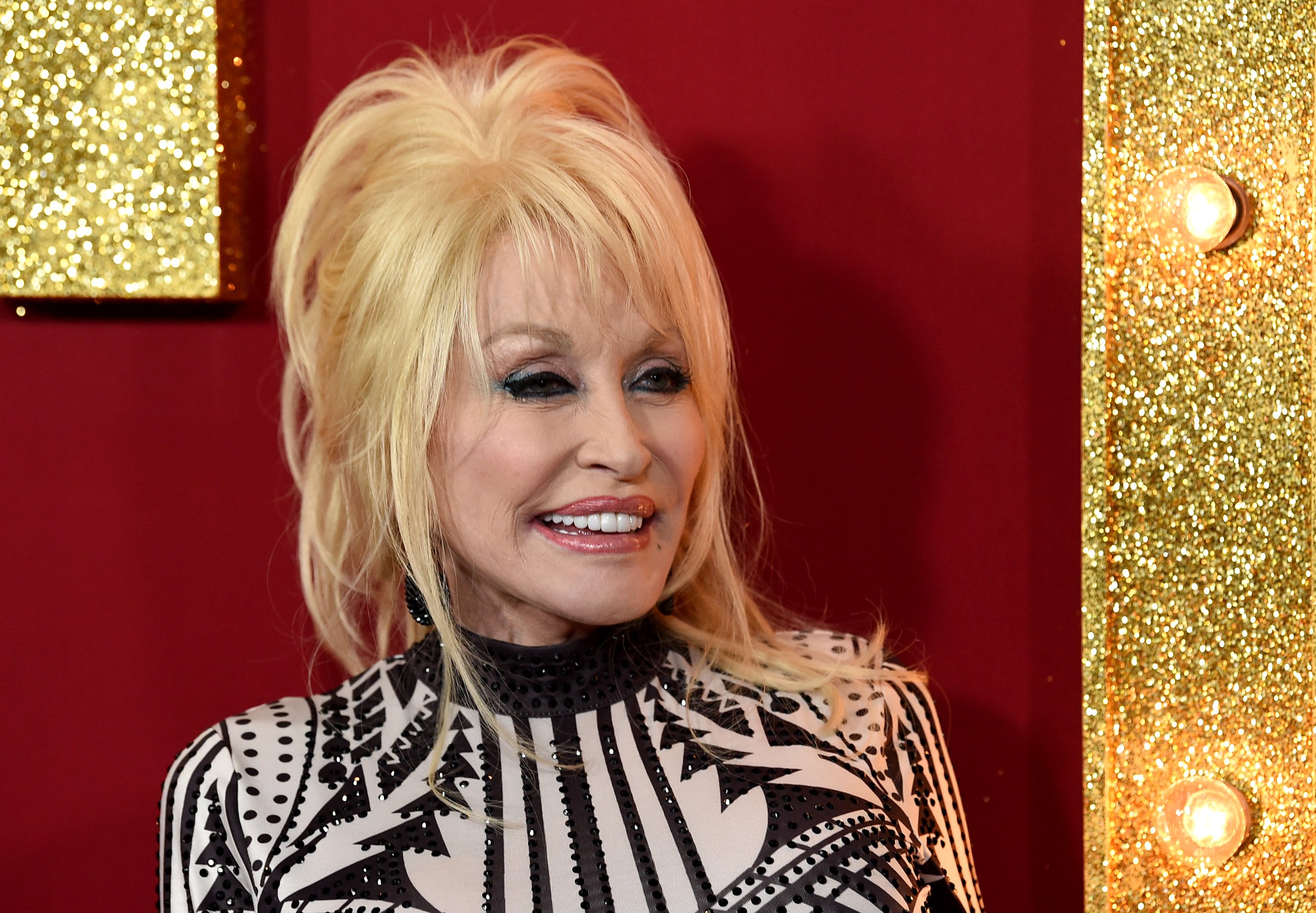 Dolly Parton at the premiere of 'Dumplin'' on the red carpet. She's in a black and white top and looking to the left of the camera, smiling.